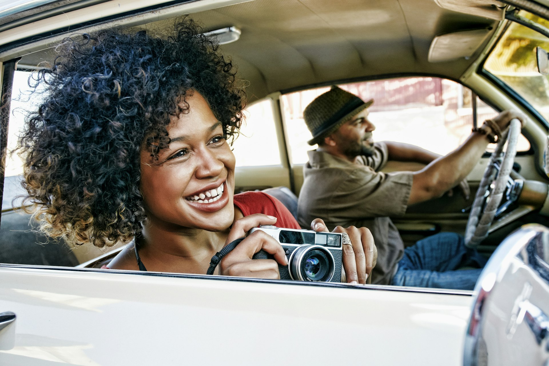 A man and woman driving in a vintage car, the woman is holding a camera and leaning toward the window