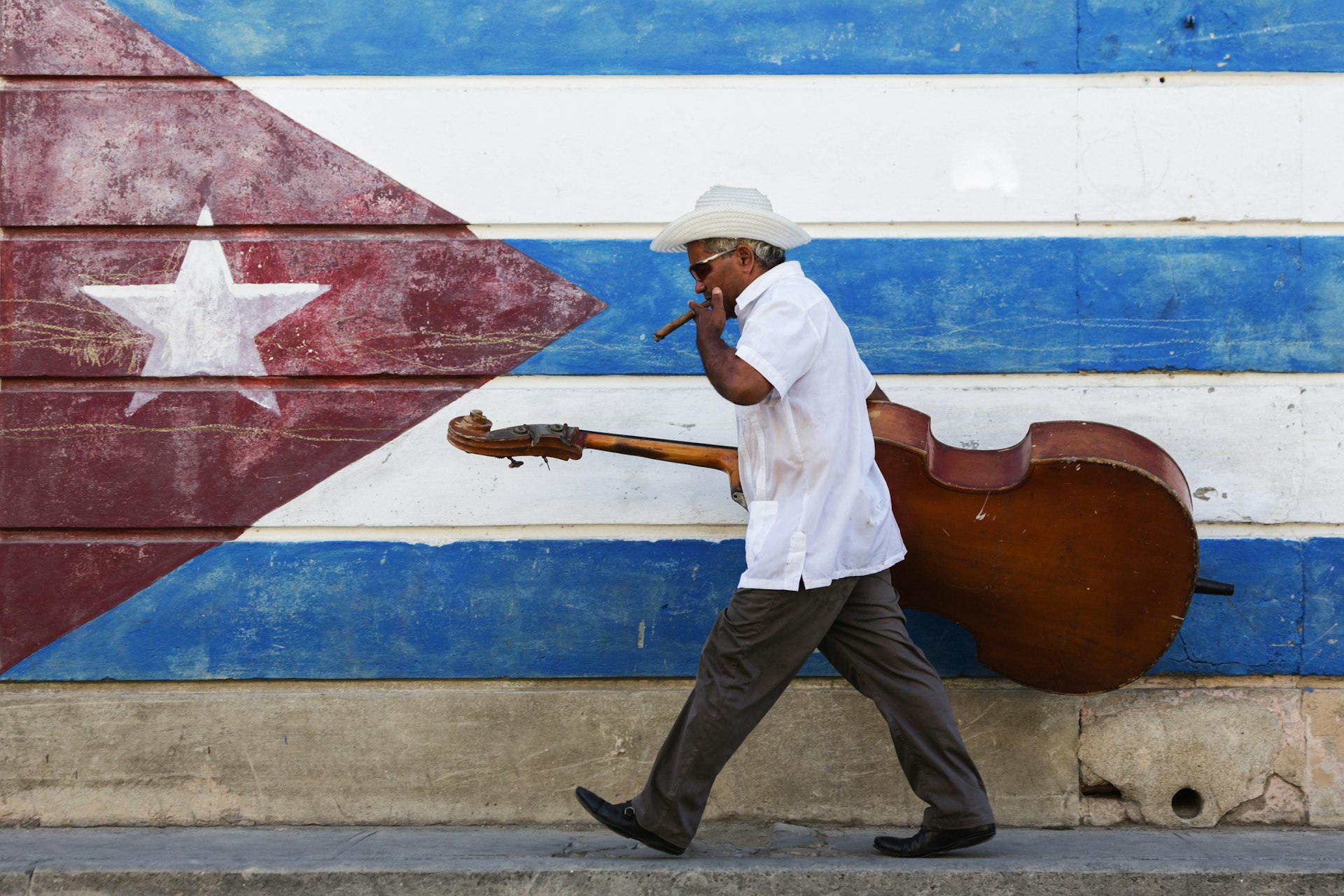 A man carrying a large bass instrument smokes a cigar as he walks by a Cuban flag painted on a wall
