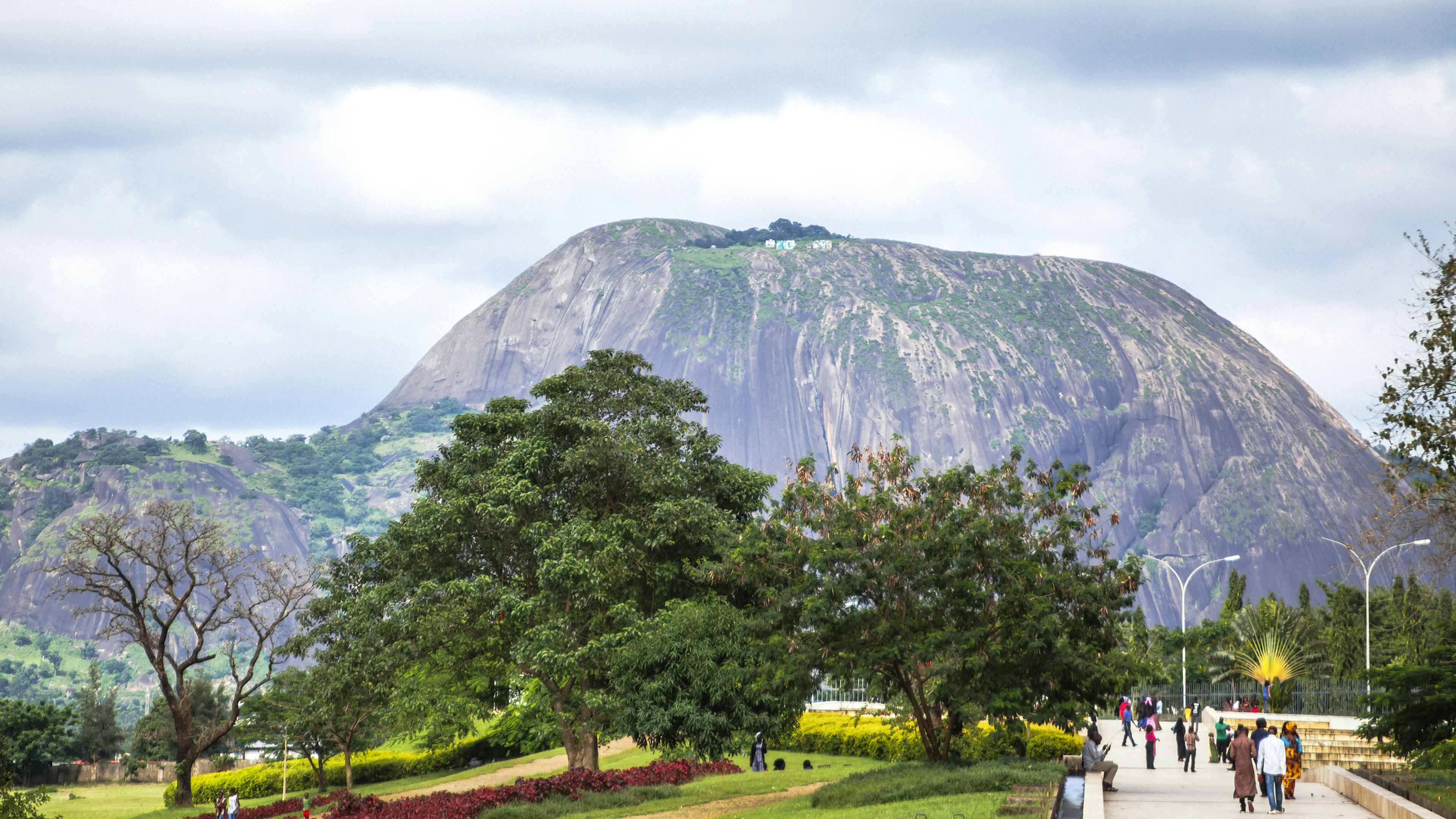 People walk the wide paths of a large parkland backed by a huge rocky hill