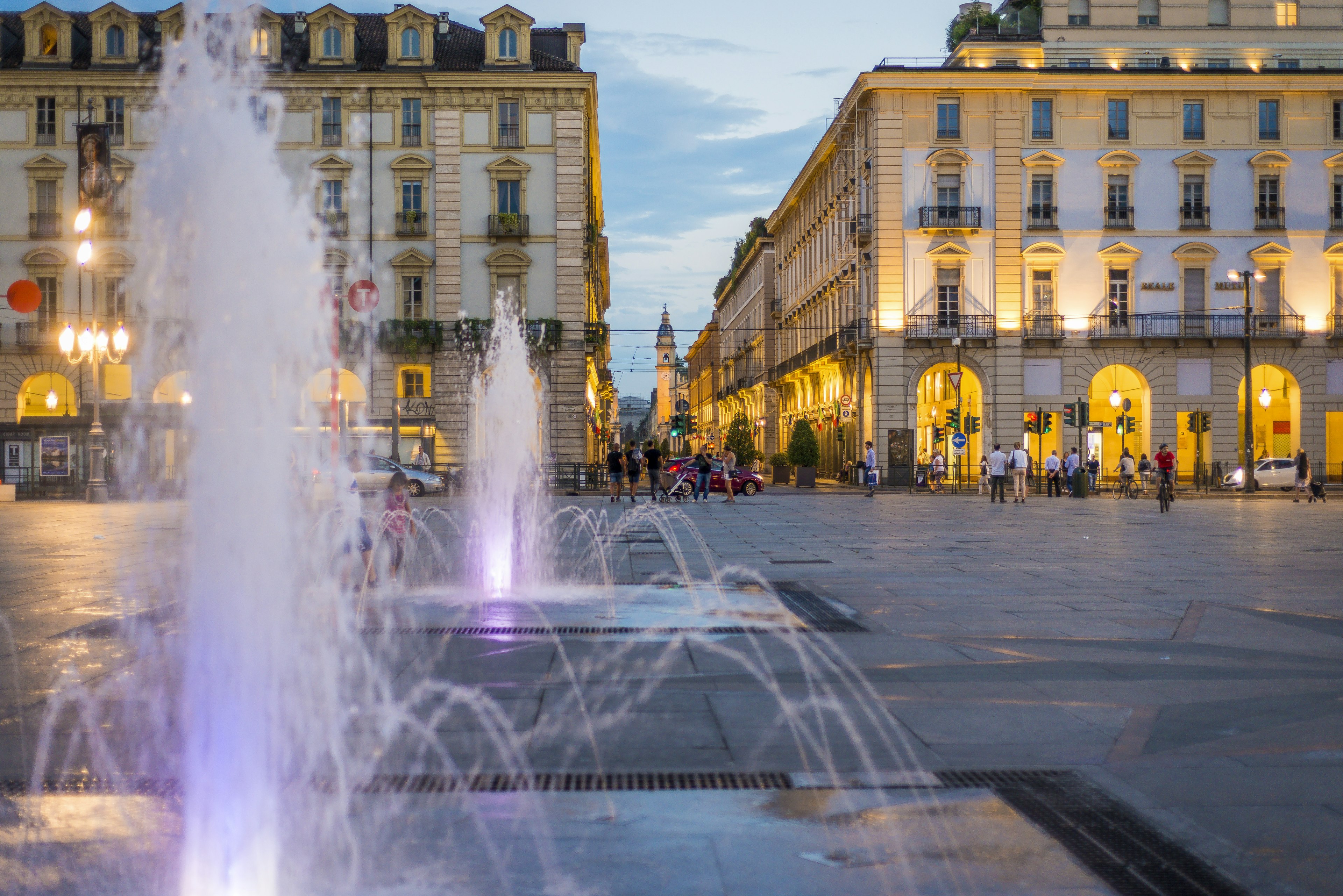 A close-up of fountains in front of people walking through porticoes of old buildings at dusk in the Piazzo Castello, Turin, Italy