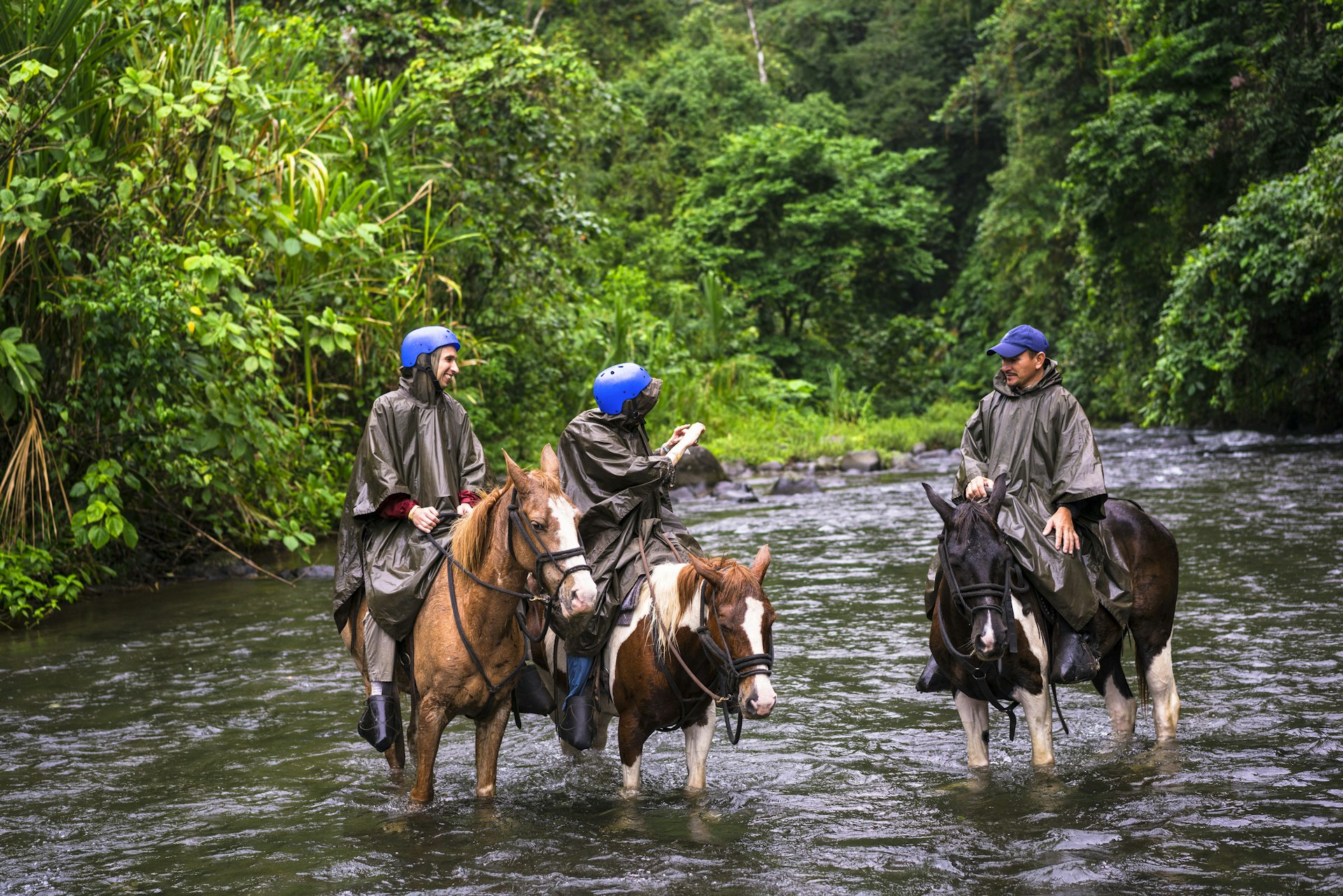 Three people on horseback wearing wet weather gear as their horses stand in a river in a jungle