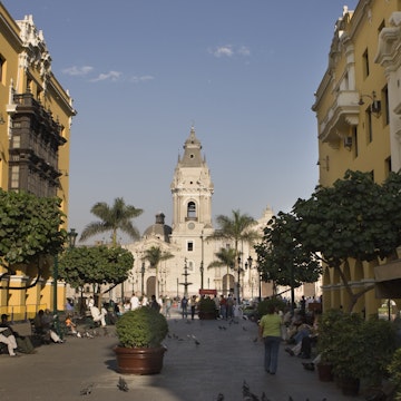 People walk down the colorful Pasaje Santa Rosa in Lima, Peru with the Municipal Palace and art gallery on the left, a restaurant on the right and the Plaza de Armas and Cathedral of Lima in the distance.
905328568