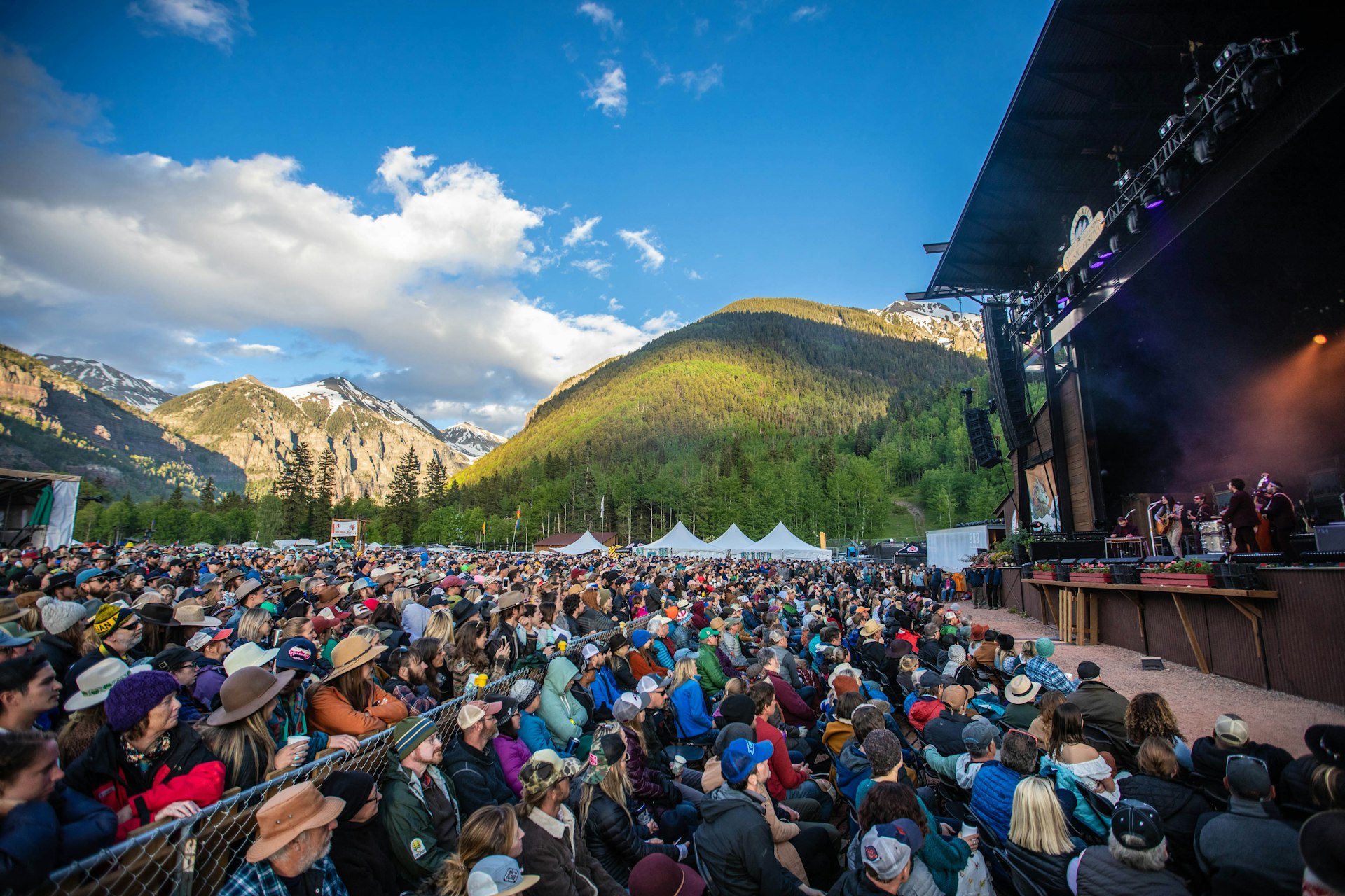 People gather at a music festival stage in front of beautiful mountains. 