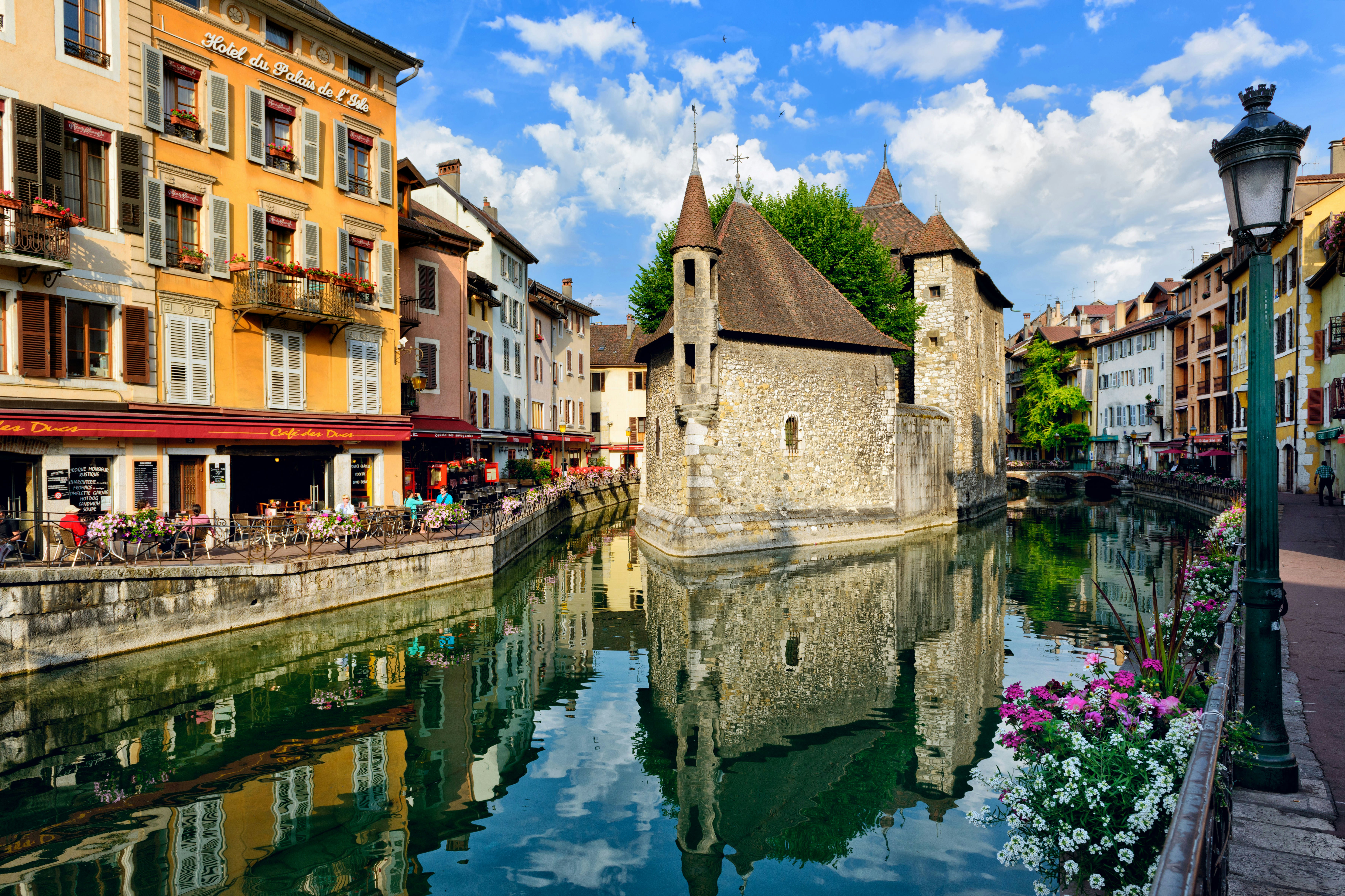 People drink coffee near the River Thiou in Old Town, encircling the medieval palace perched mid-river - the Palais de l'Isle in Annecy, France.