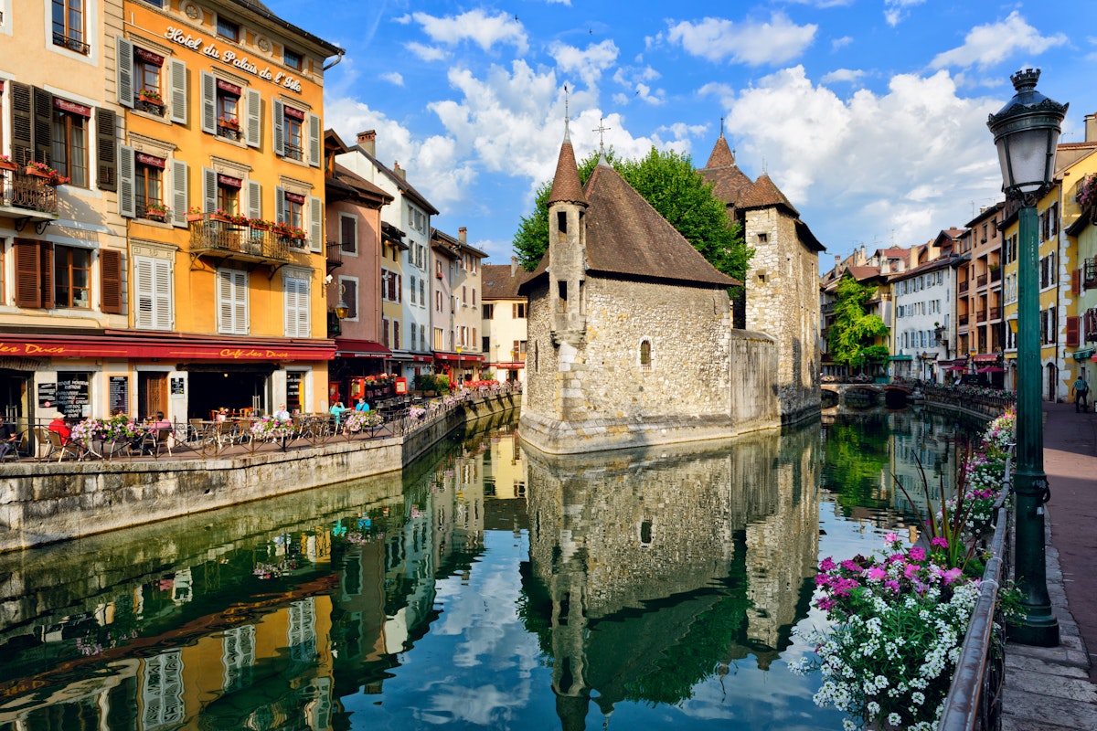 People drink coffee near the River Thiou in Old Town, encircling the medieval palace perched mid-river - the Palais de l'Isle in Annecy, France.