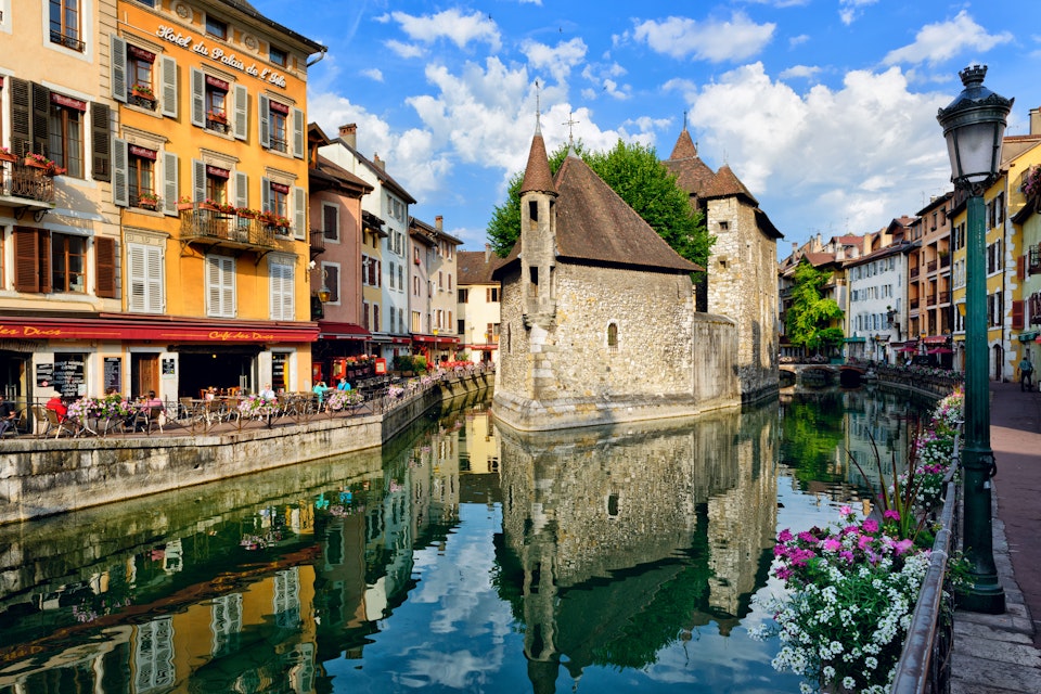 People drink coffee near the River Thiou in Old Town, encircling the medieval palace perched mid-river - the Palais de l'Isle in Annecy, France.