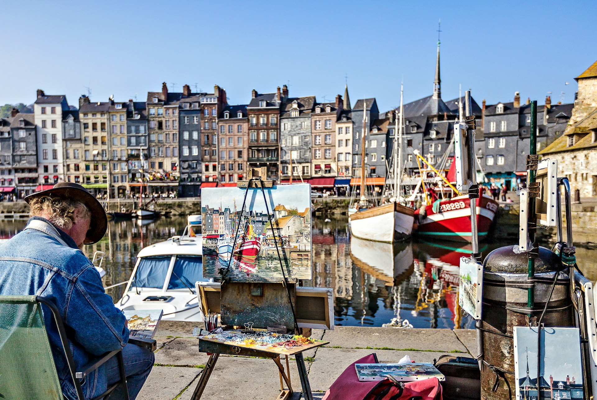 An artist paints at an easel in the historic, sailboat-filled harbor of Honfleur, Normandy, France