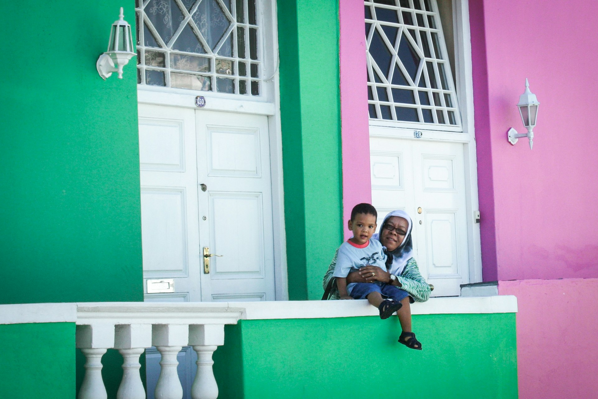 A woman stands with a child who is sat on a wall against a colorful pink and green building