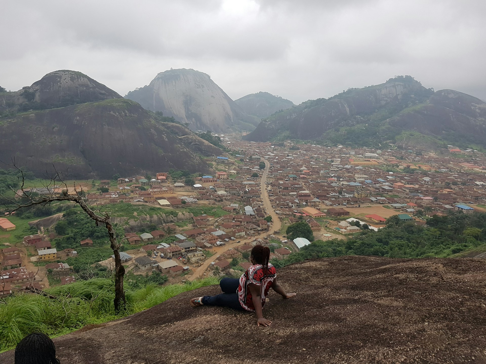 A woman sits on the edge of a viewpoint looking out to the red roofs of the settelments below the surrounding hills 