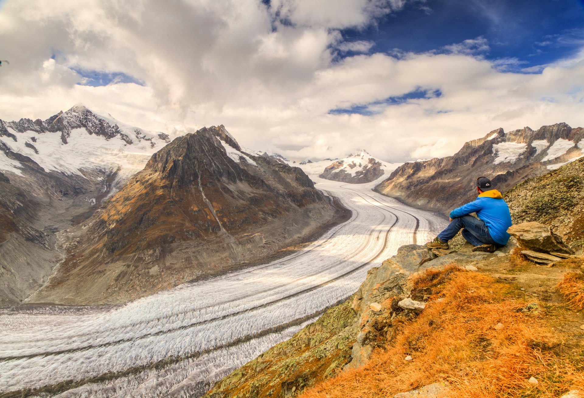 A hiker sits down and takes in the vast Aletsch Glacier as it snakes around the mountains in the Alps