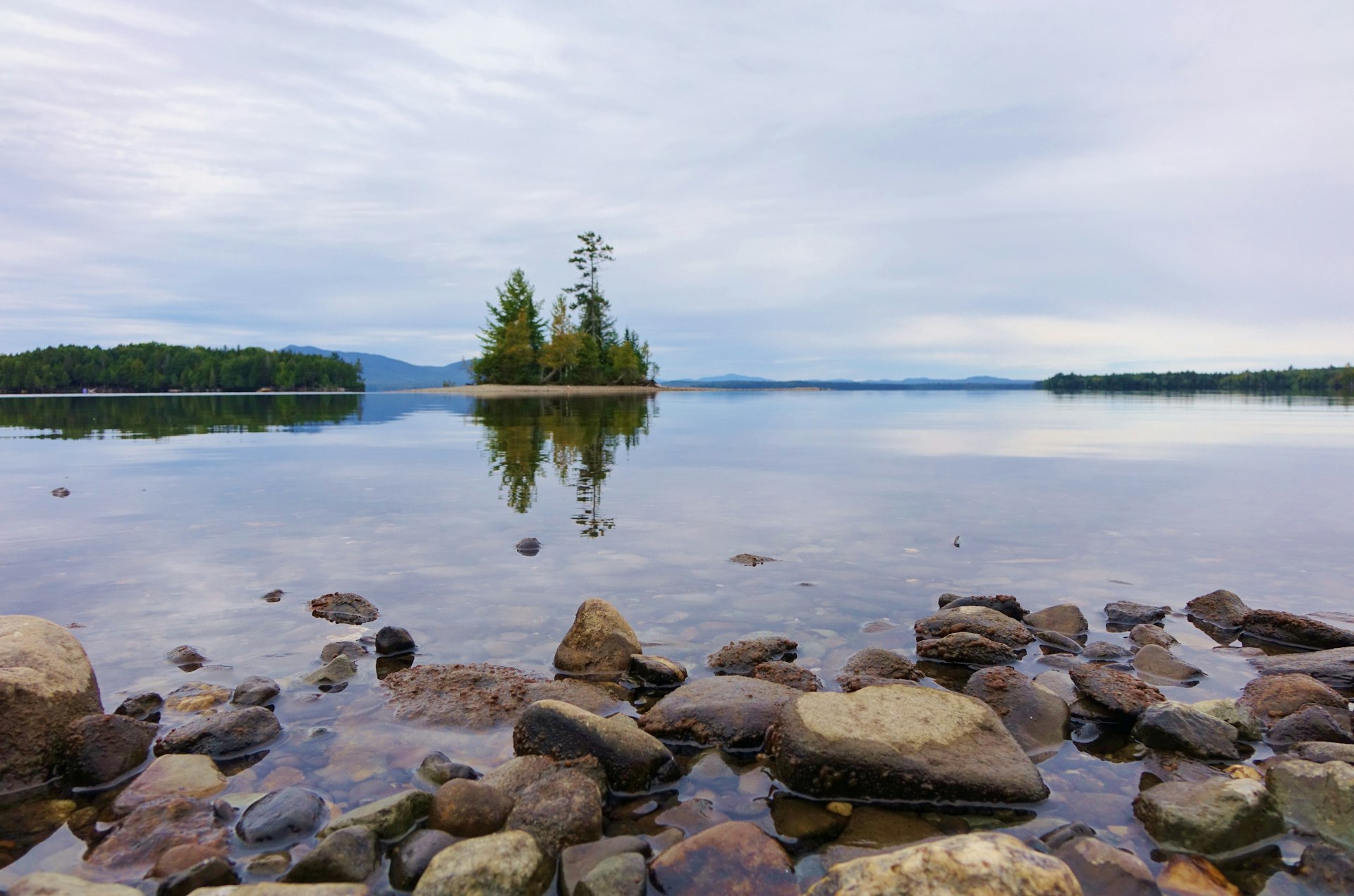 From ground level on a rocky beach on Moosehead Lake in Lily Bay State Park in Maine you see a very calm lake with trees on a small island reflected on the still water under a cloudy autumn sky