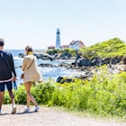 Cape Elizabeth, USA - June 10, 2017: Young couple walking on trail by edge of cliff rocks at Portland Head Lighthouse in Fort Williams park in Maine during summer day