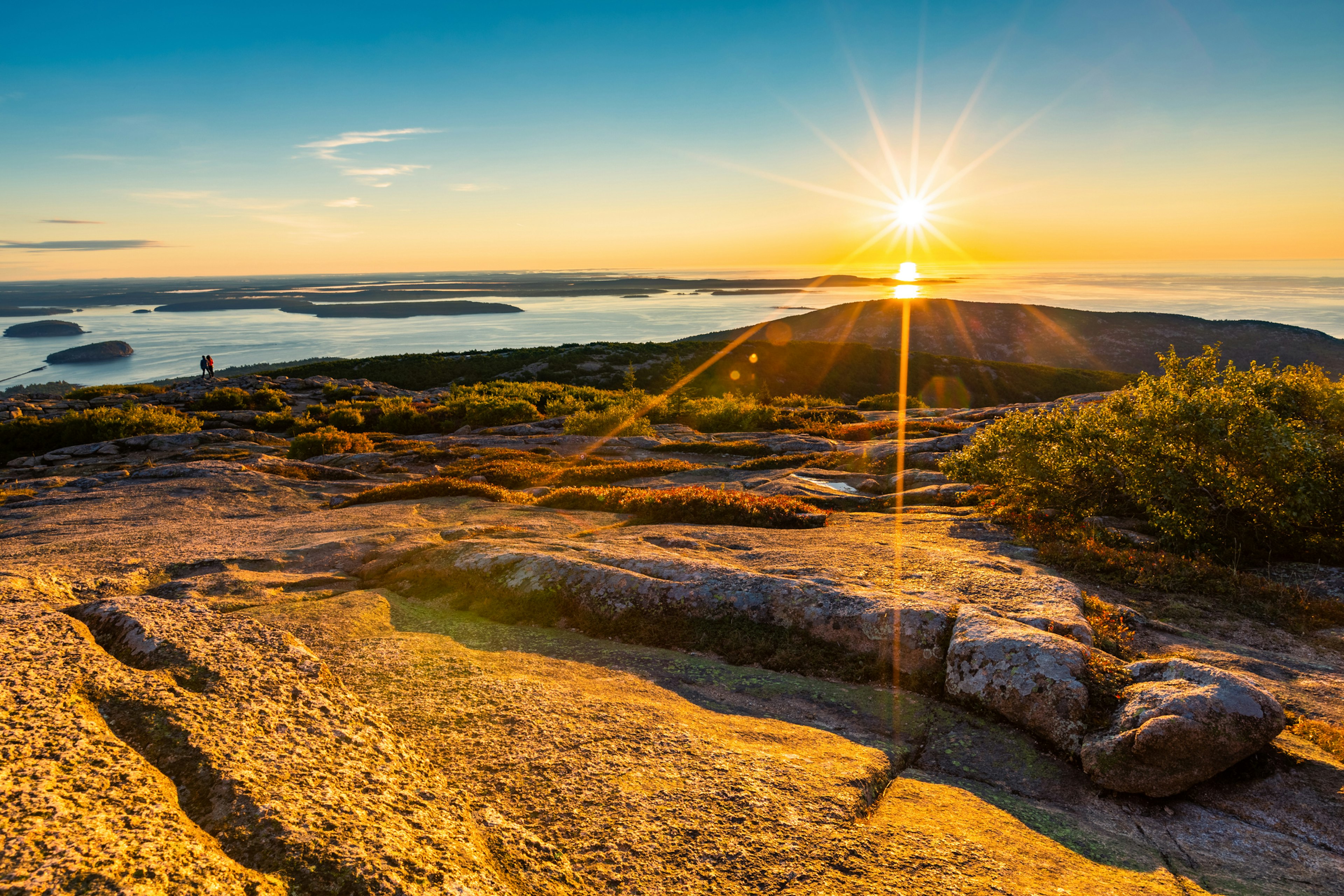 Two people walk along the edge of a mountain ridge as the sun rises in the sky casting an orange glow over the surroundings