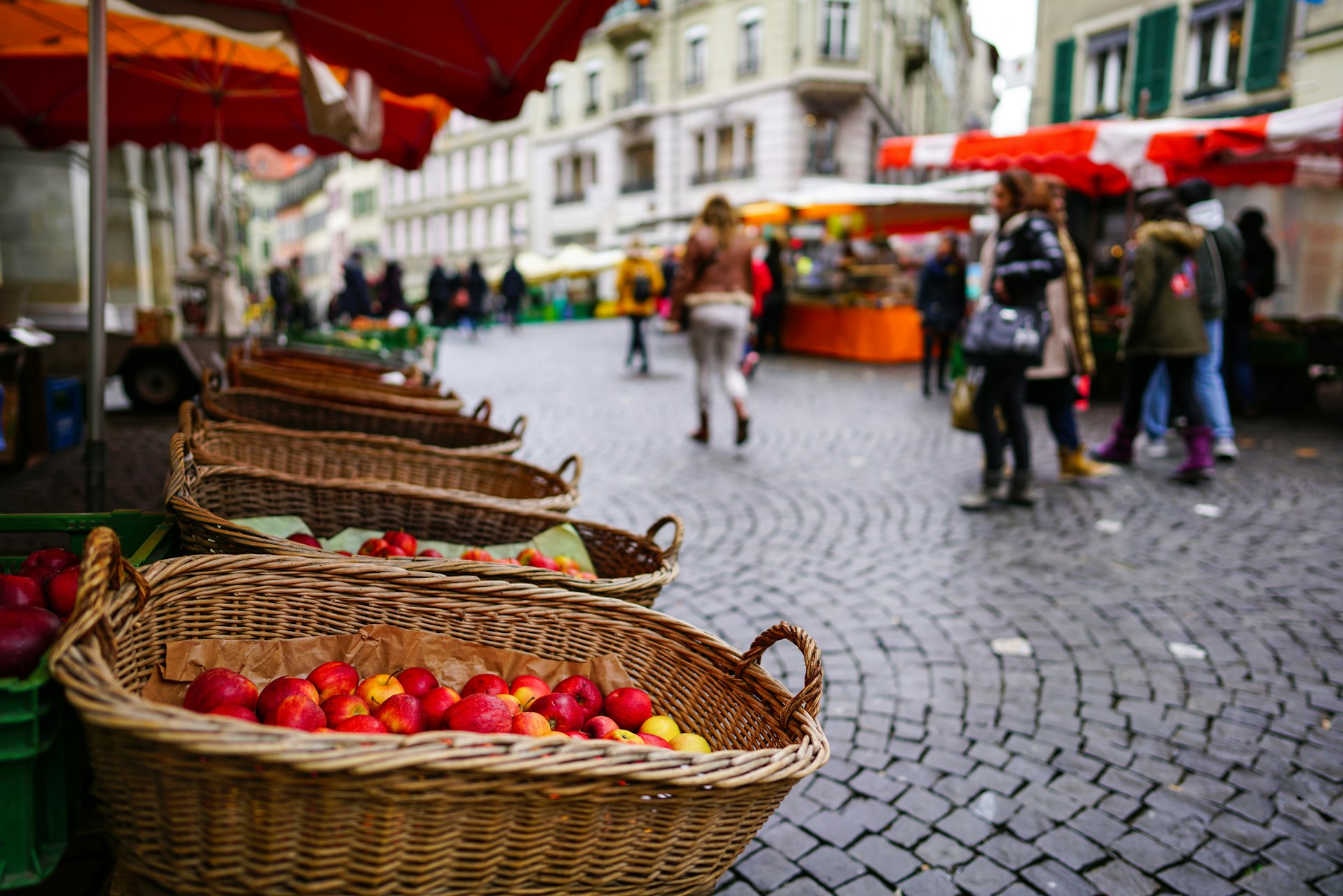 A fruit and veg stall in a cobbled city square on market day as people walk by