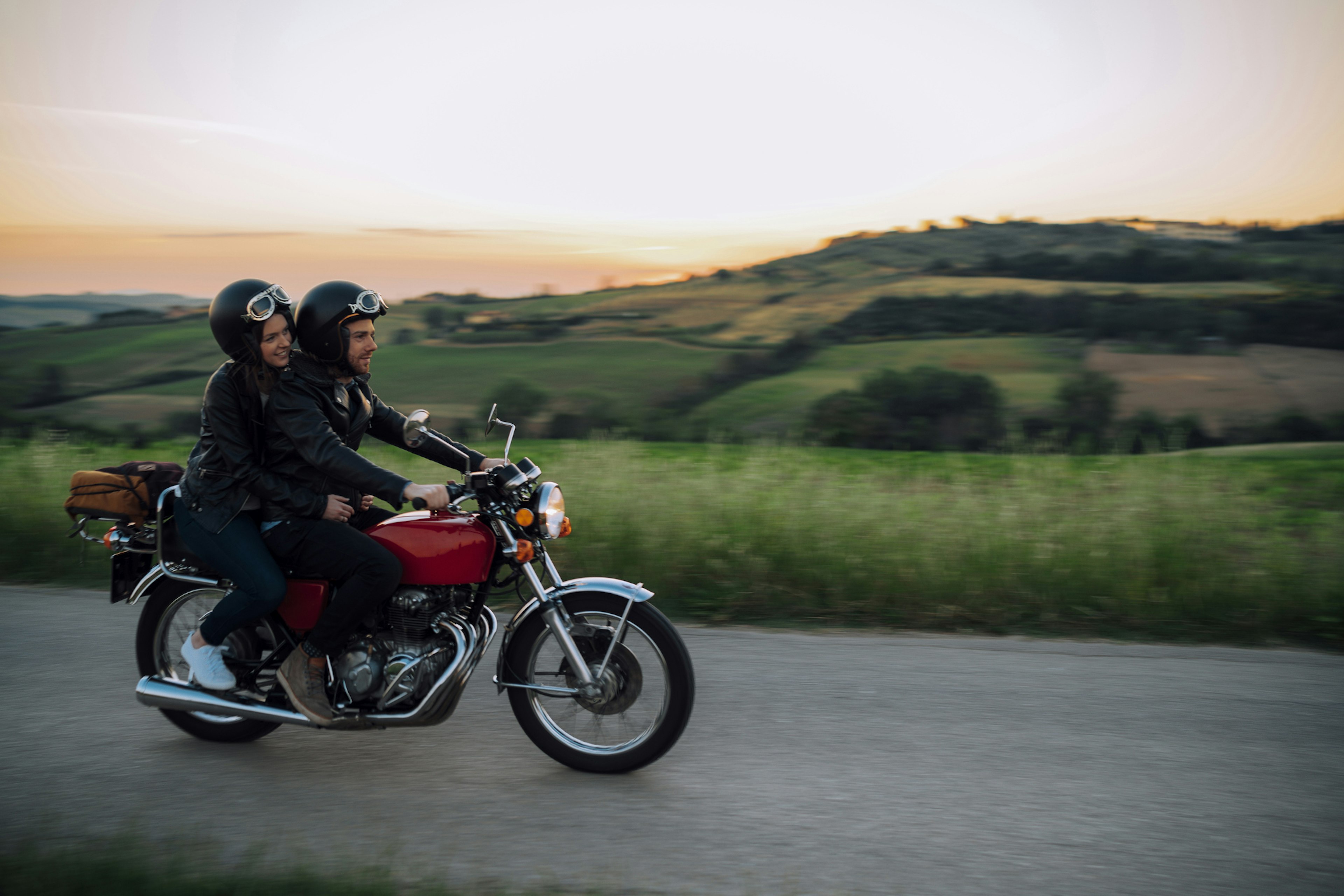 A male and female couple ride on the back of a motorbike through the Tuscan countryside as the sun sets