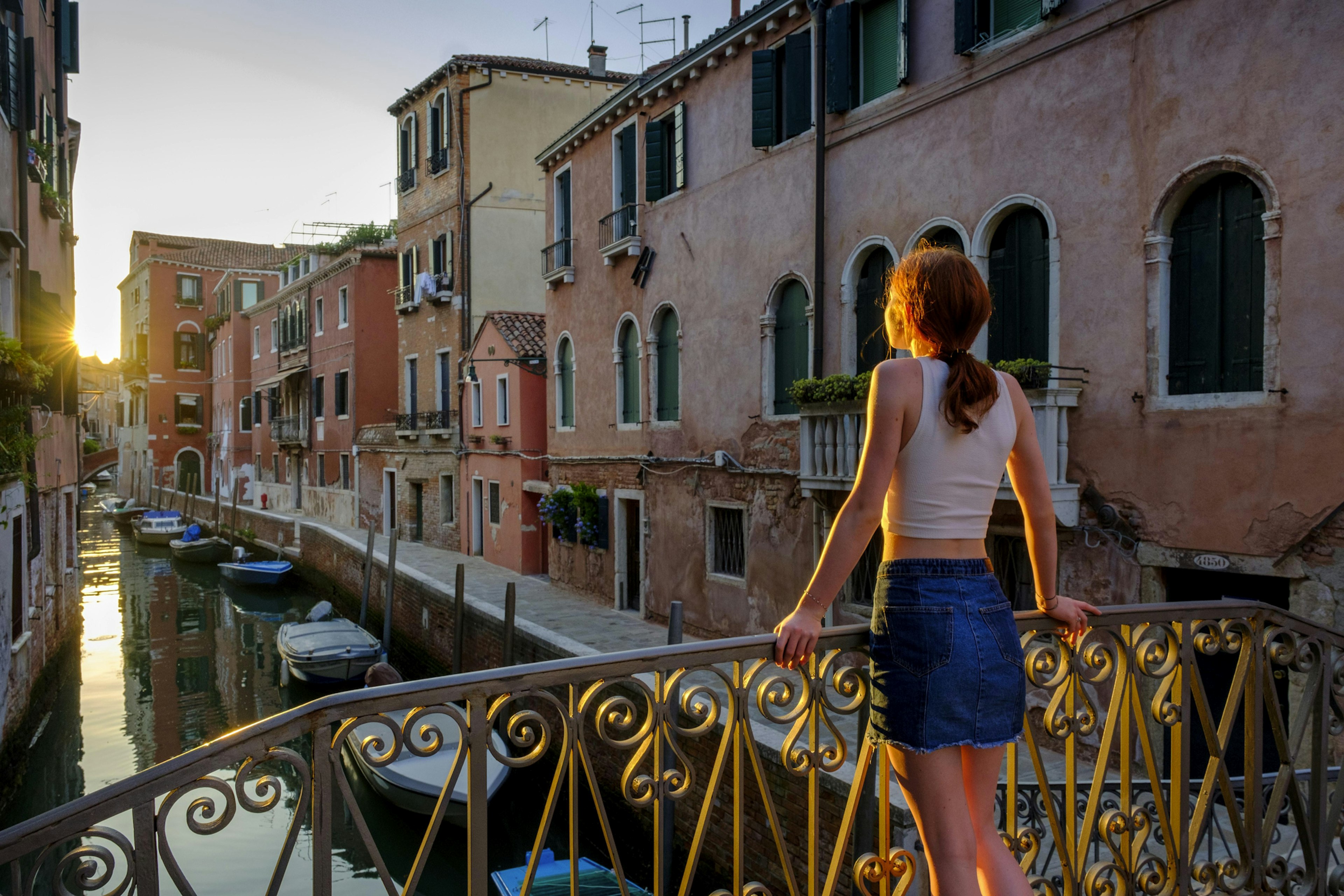 A ginger white woman seen from behind, stands on a bridge overlooking a canal Young woman leaning against a bridge on the Rio Santa Catherina in the evening light, Cannaregio, Venice,