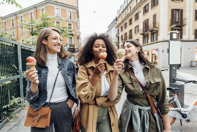 Three smiling women eating ice cream while walking in an Italian city and laughing together