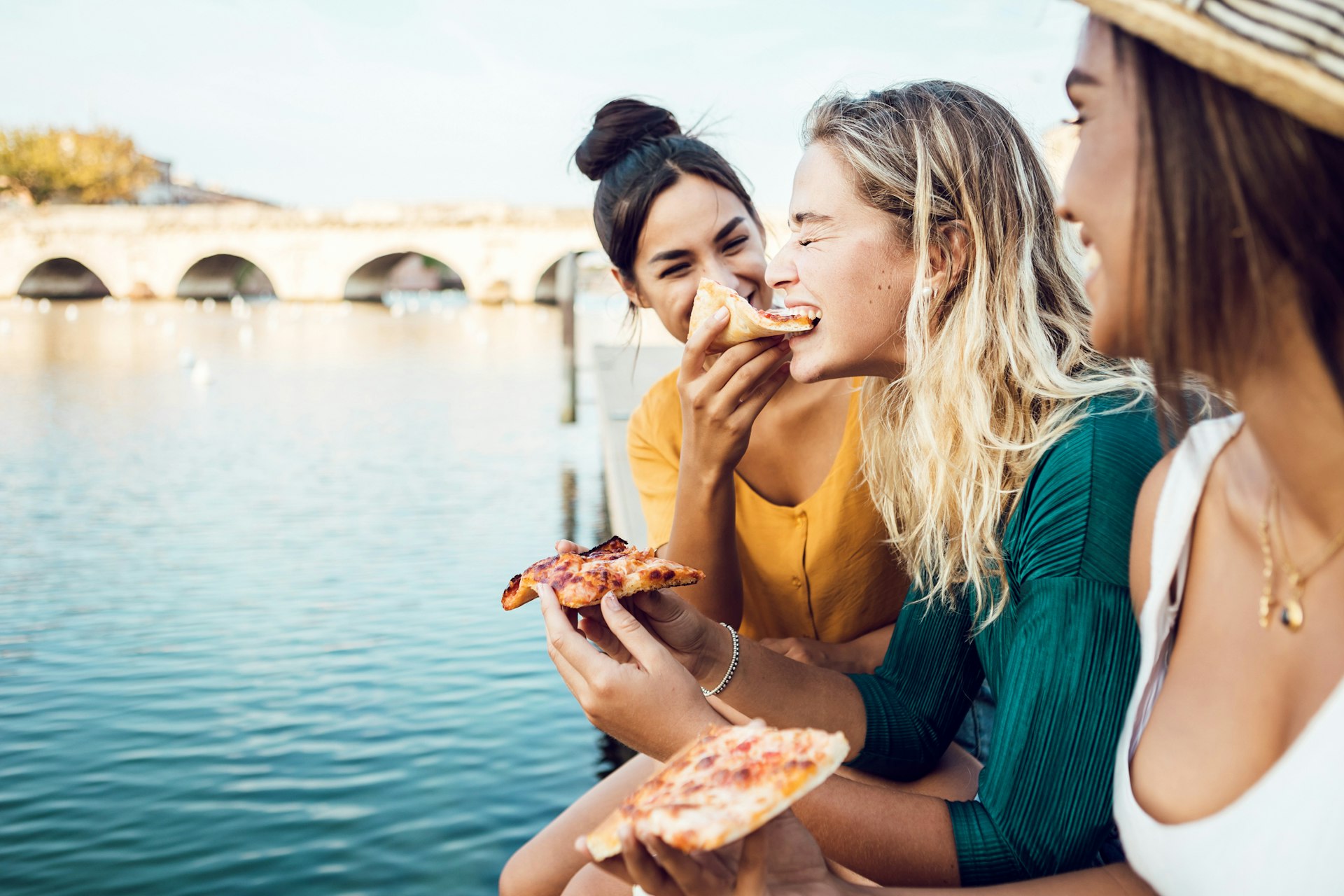 Three friends sit by a river in Italy eating pizza with cheese and tomato