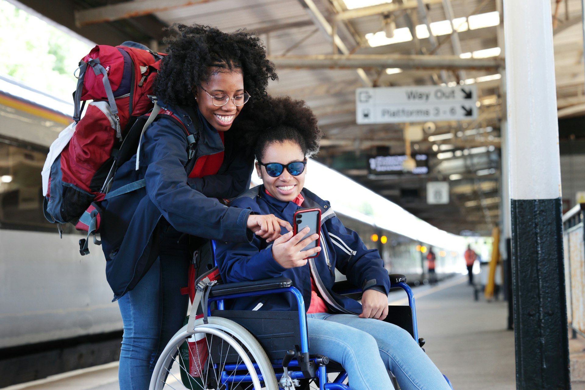 Two women, one in a wheelchair and one standing, look at a smartphone on a train platform
