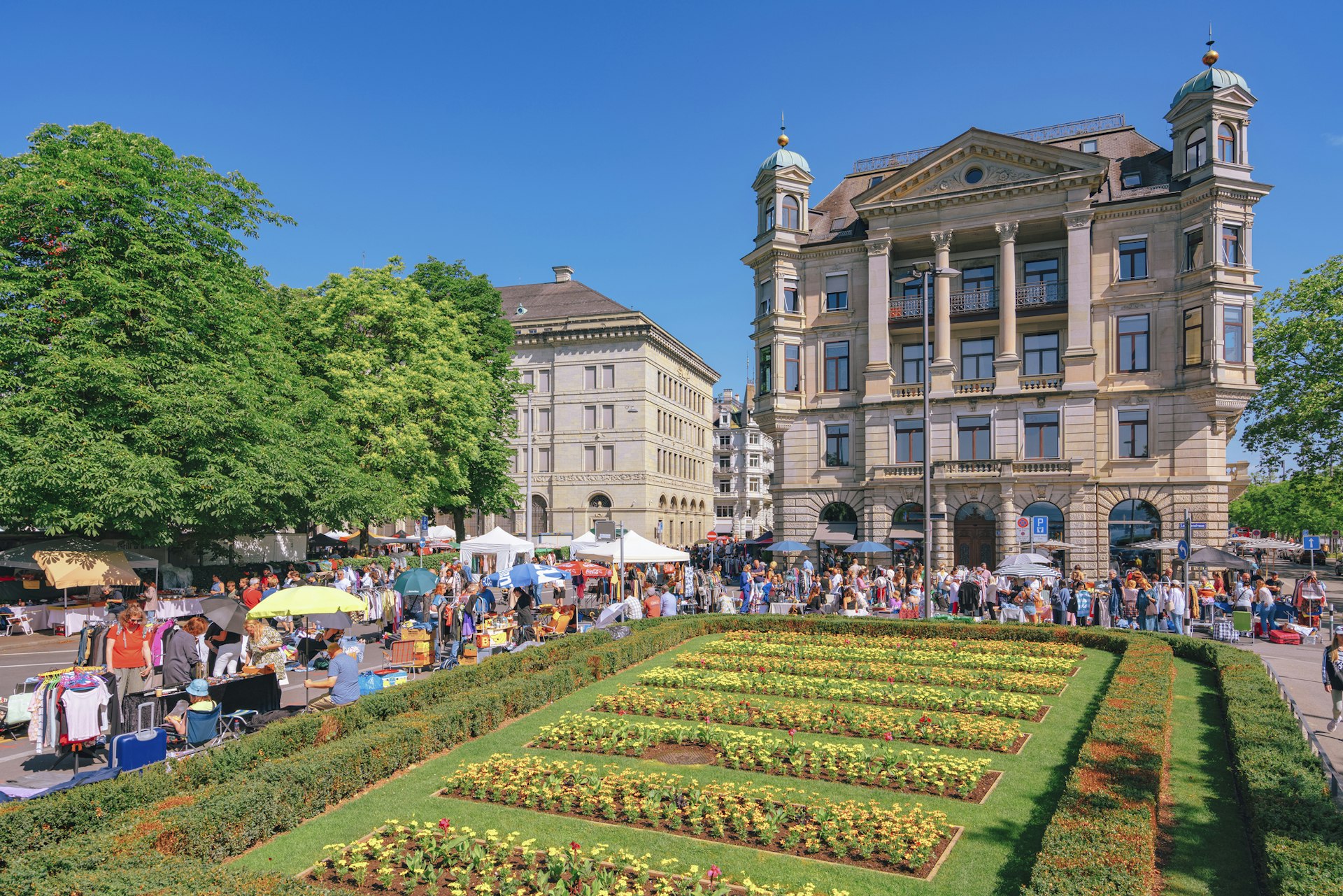 A neat flower garden in front of a grand building is surrounded by market stalls stocked with bric-a-brac for sale