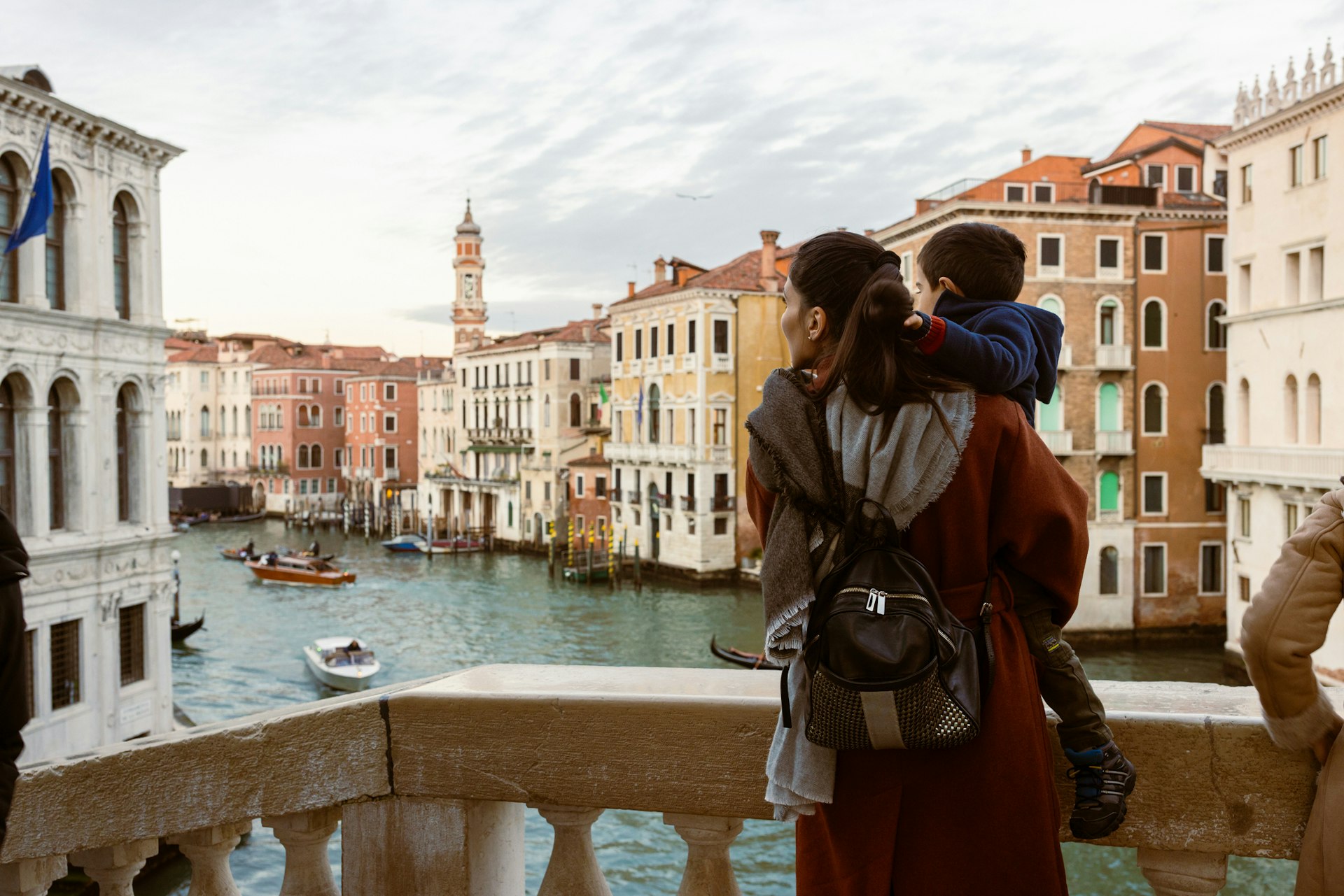 Mother is carrying her son as they enjoy a tourist visit to Venice. They are standing on a bridge overlooking the Grand Canal, with gondolas and boats passing by. The magnificent architecture and stunning waterways of Venice are on full display, providing a picturesque backdrop for their memorable trip