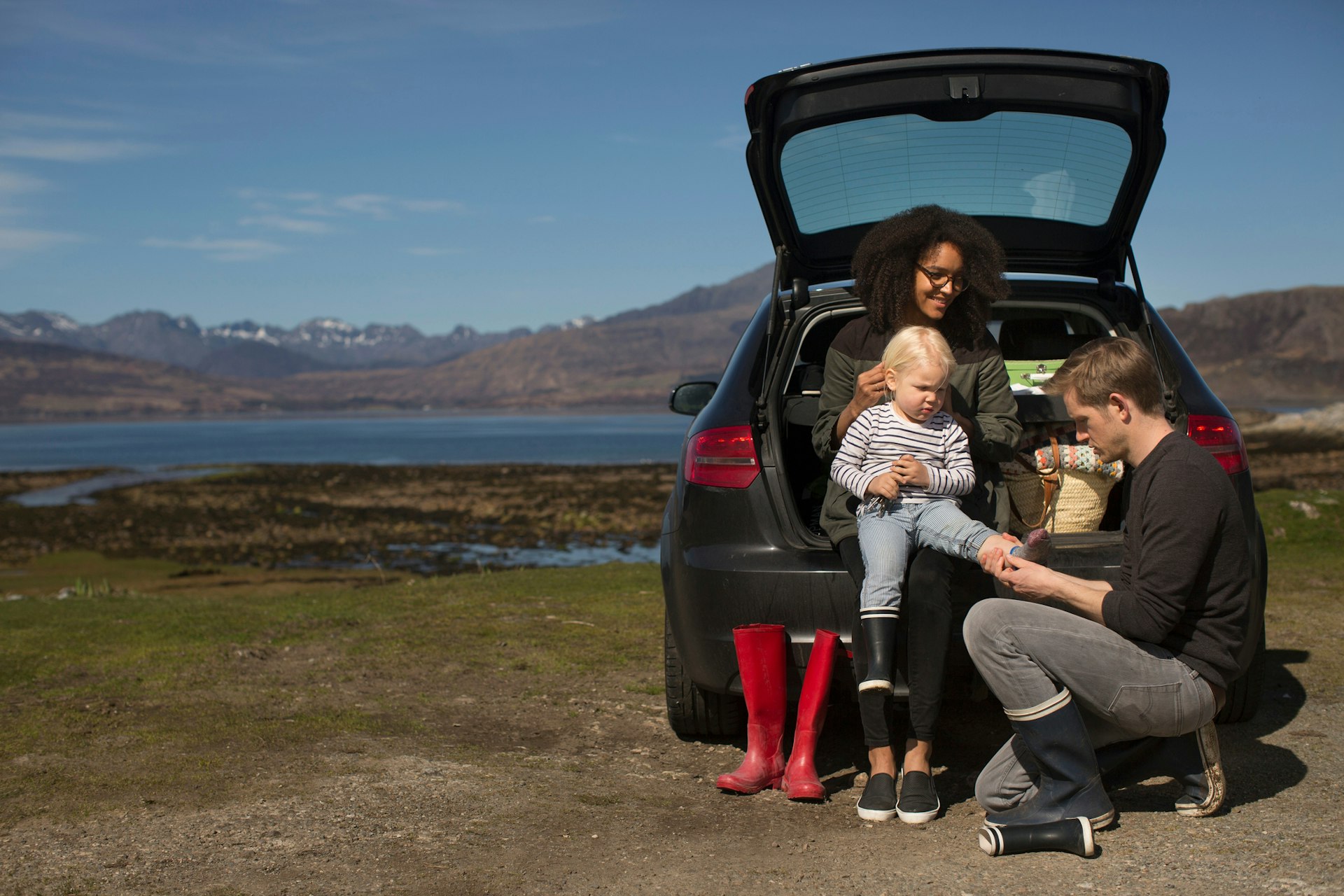 A family of three sit on the open trunk of a car parked by a lake changing shoes following a paddle