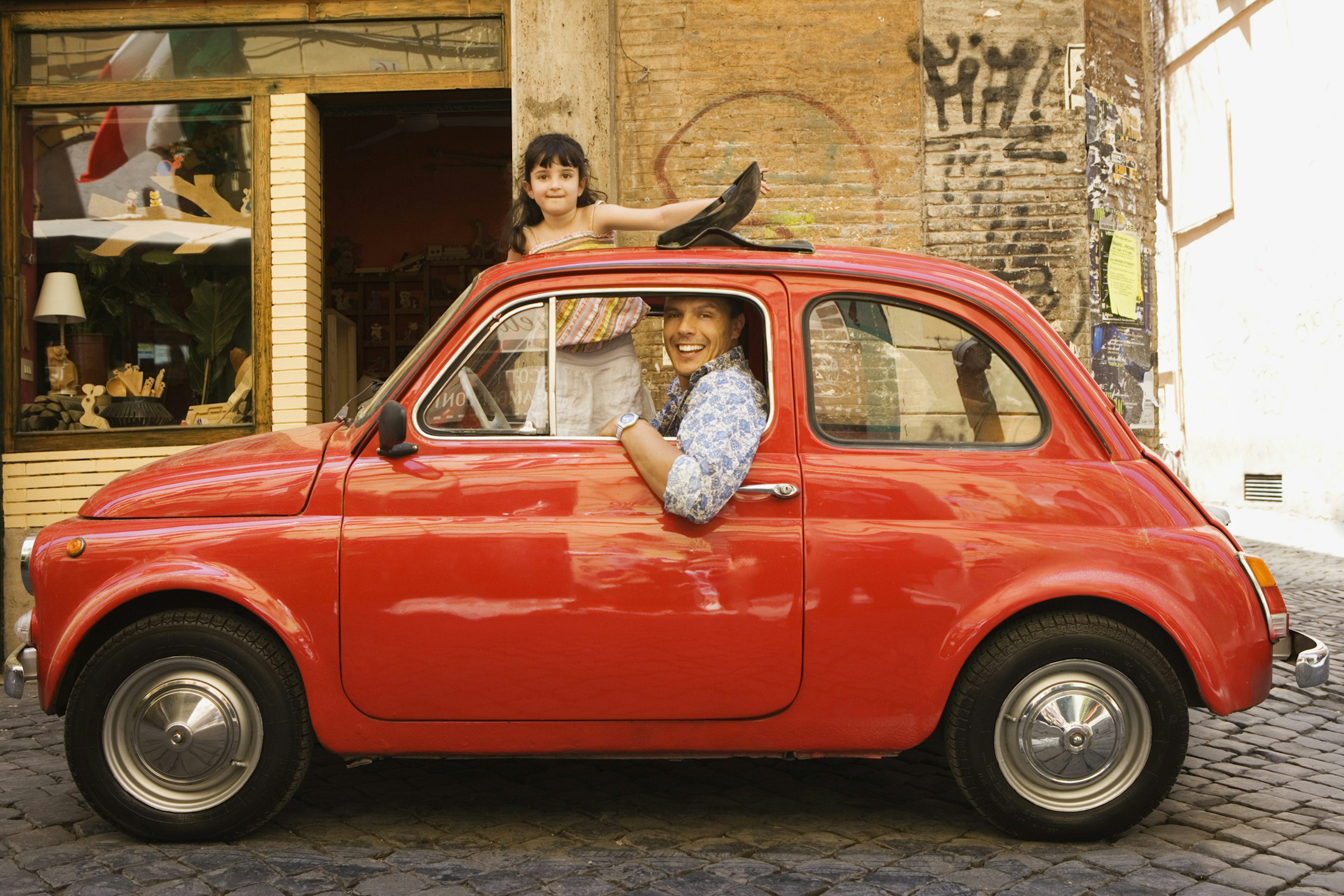 A smiling father and daughter in a Fiat 500 car in Italy, with the daughter standing up outside the sunroof