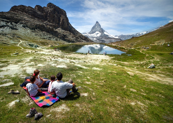 Summertime in Switzerland. A family of four is having fun relaxing on the lawn in front of the Matterhorn. The mountain peak is reflecting into the water of a mountain lake. Horizontal composition, Outdoor experience.
915075652