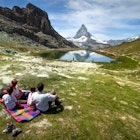 Summertime in Switzerland. A family of four is having fun relaxing on the lawn in front of the Matterhorn. The mountain peak is reflecting into the water of a mountain lake. Horizontal composition, Outdoor experience.
915075652