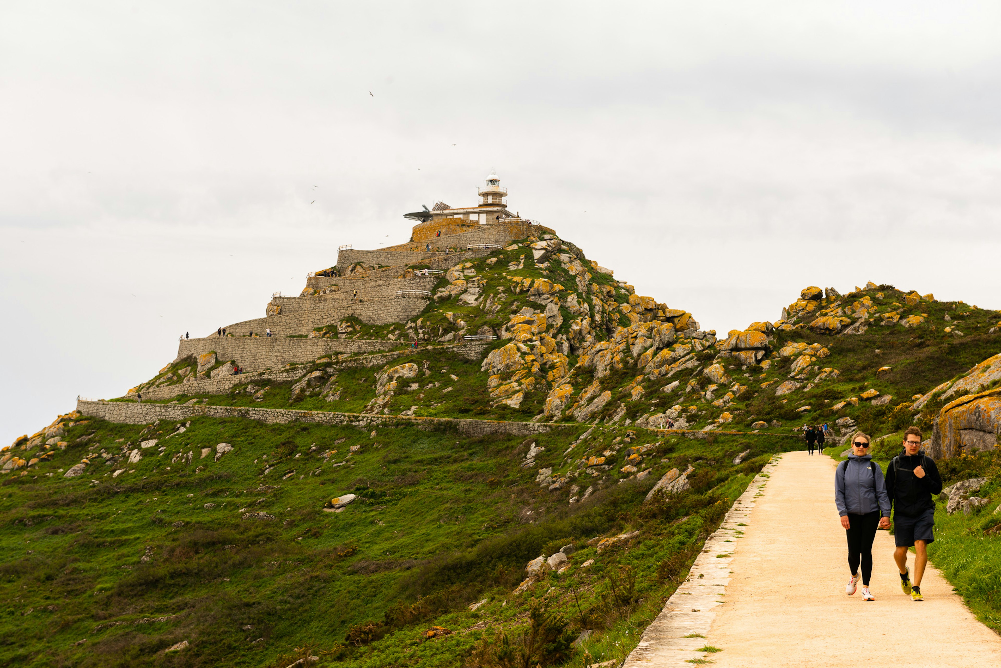 Two people walk down a path on an island leading away from a lighthouse on a clifftop