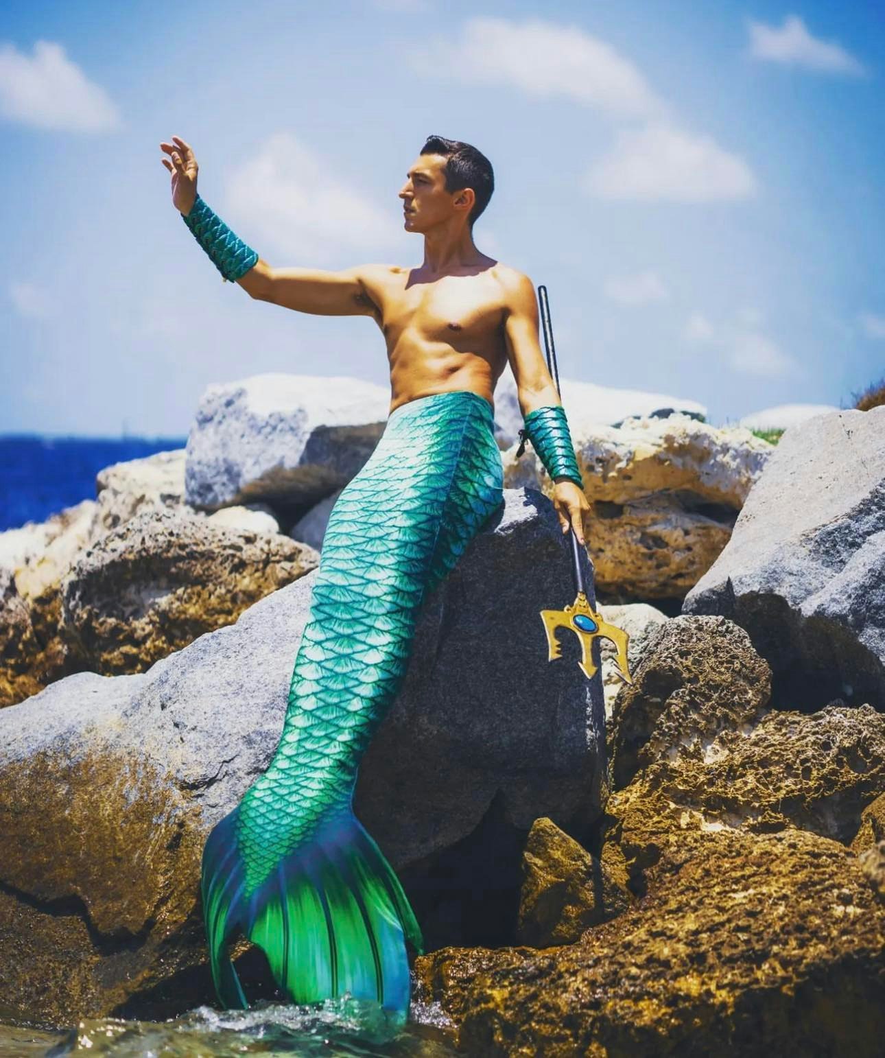 Merman Andrew sits on a rock, right arm aloft, with a three-pronged trident in his hand and his green mermaid tail drooping down towards the beach