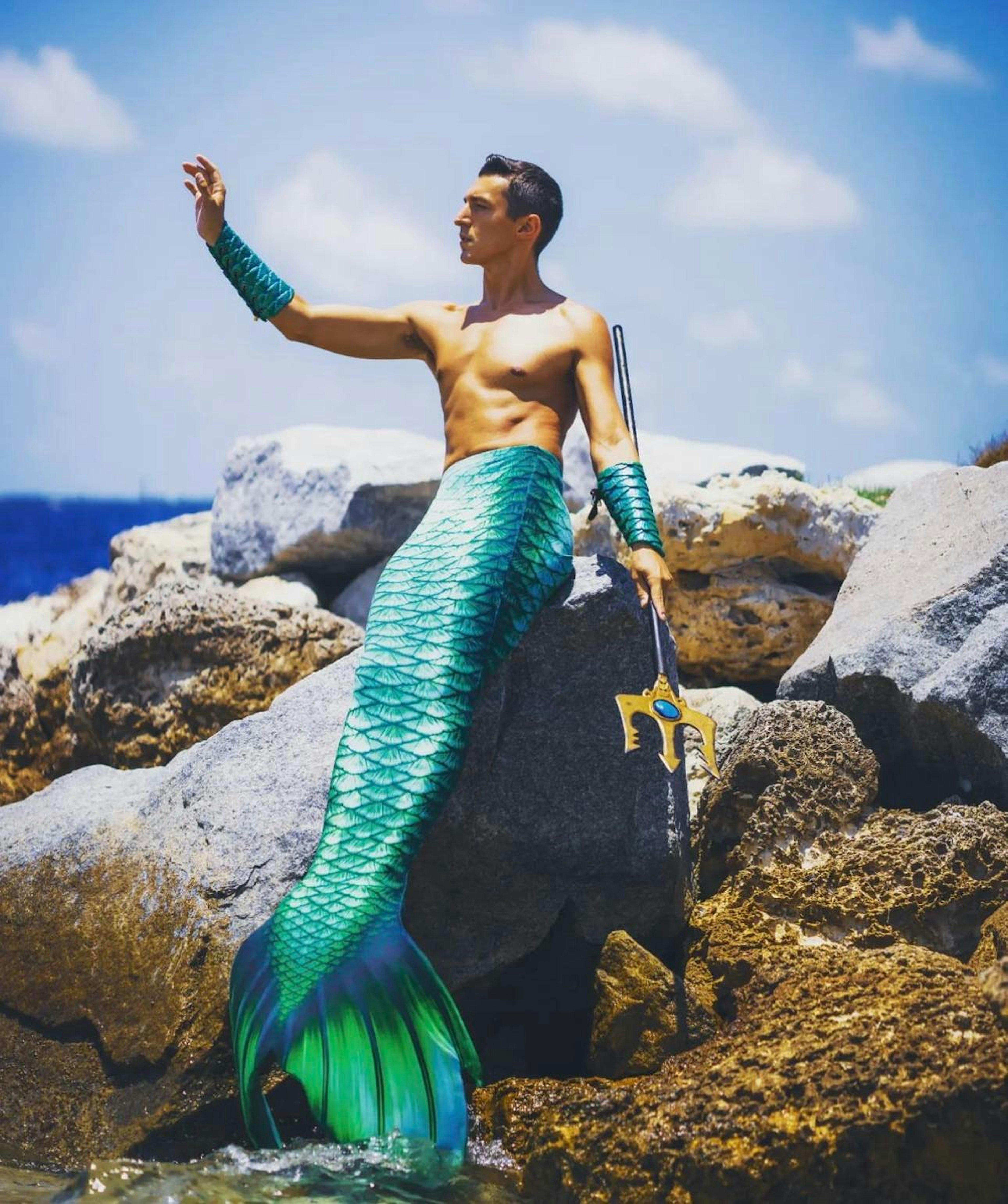 Merman Andrew sits on a rock, right arm aloft, with a three-pronged trident in his hand and his green mermaid tail drooping down towards the beach