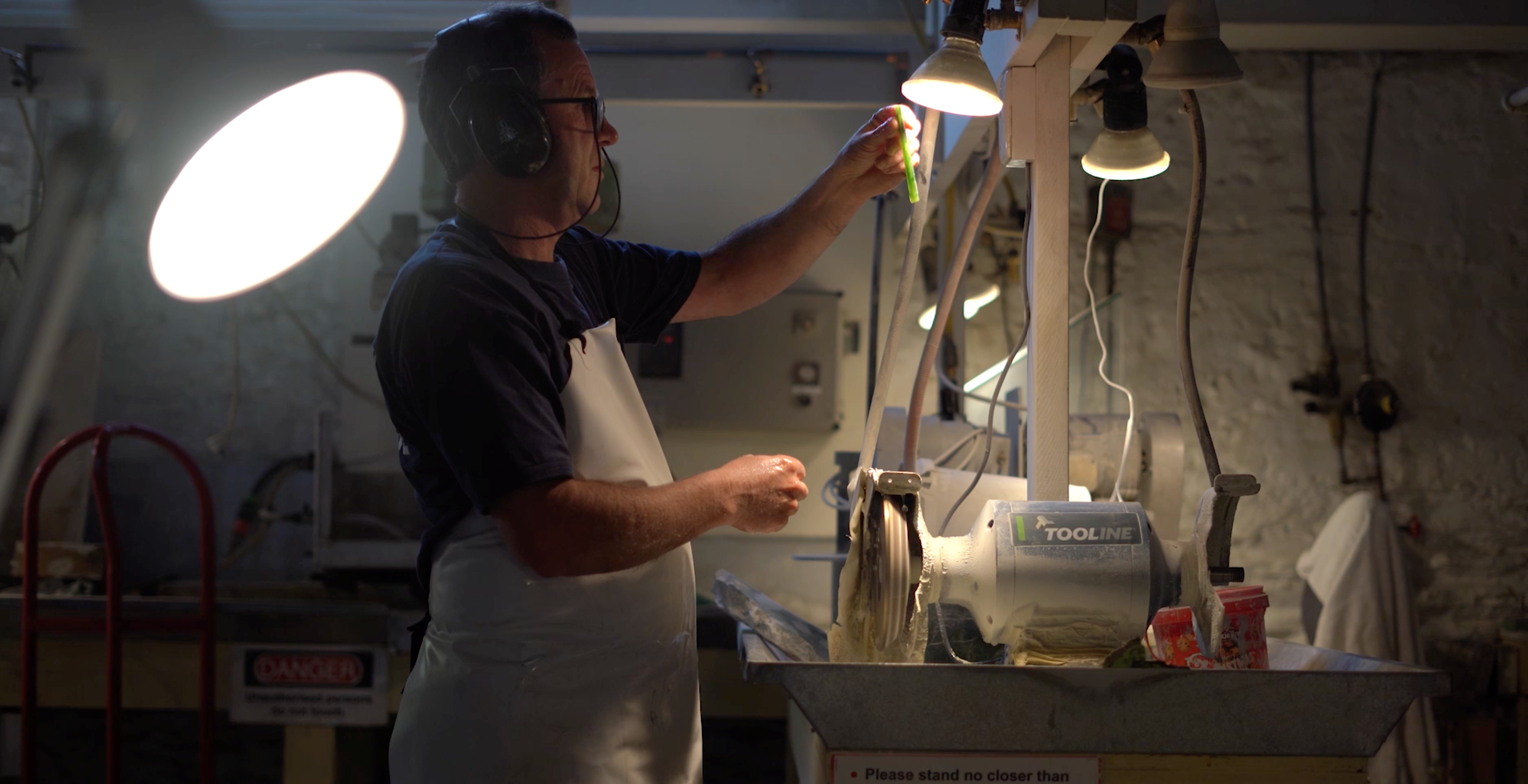 A craftsperson in a workshop holds a piece of green rock up to a light ahead of carving it