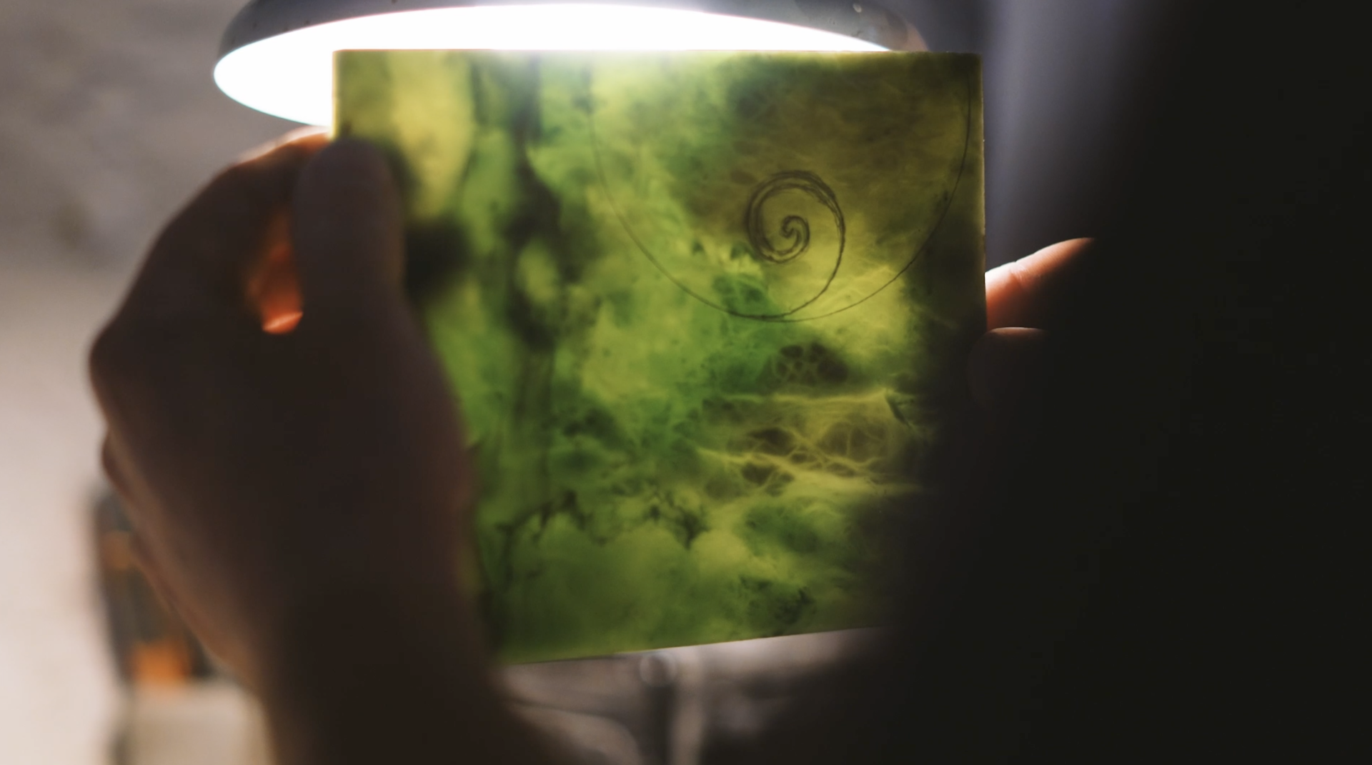 Two hands hold a piece of green stone up towards the light. A swirl shape has been drawn on