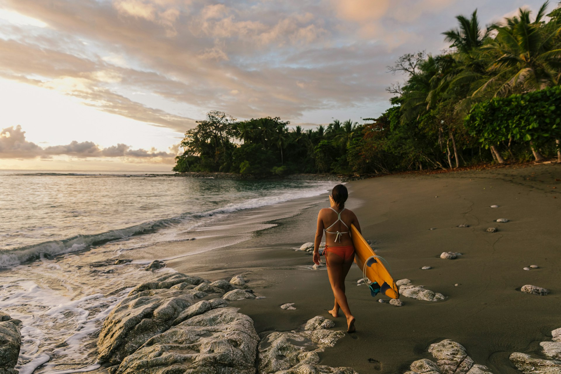 A woman walks across a beach in Costa Rica at sunset with a surfboard in her hand
