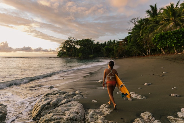 A woman walks across a beach in Costa Rica at sunset with a surfboard in her hand