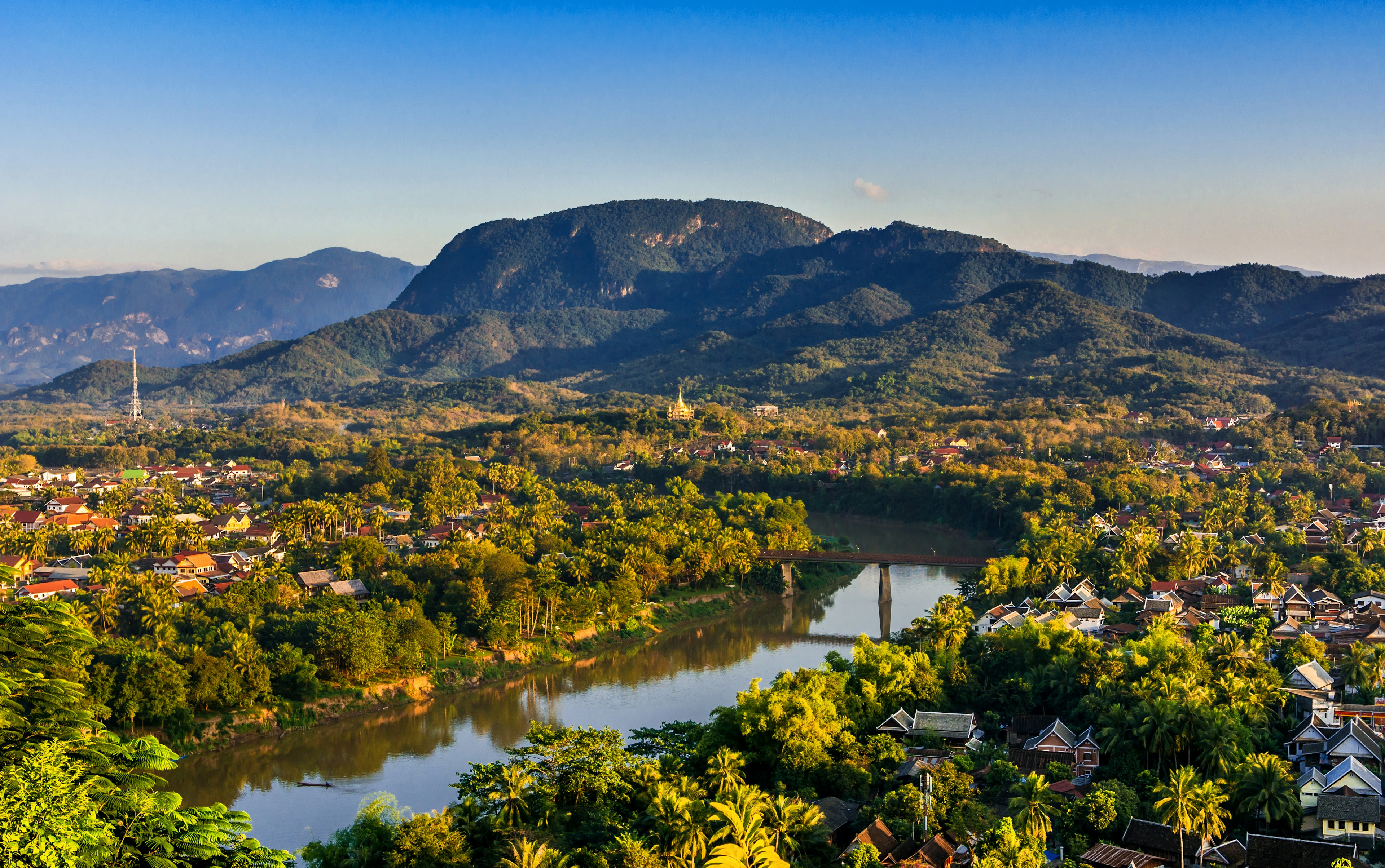 Sunset over a riverside city with dense foliage, and a range of hills rising in the distance