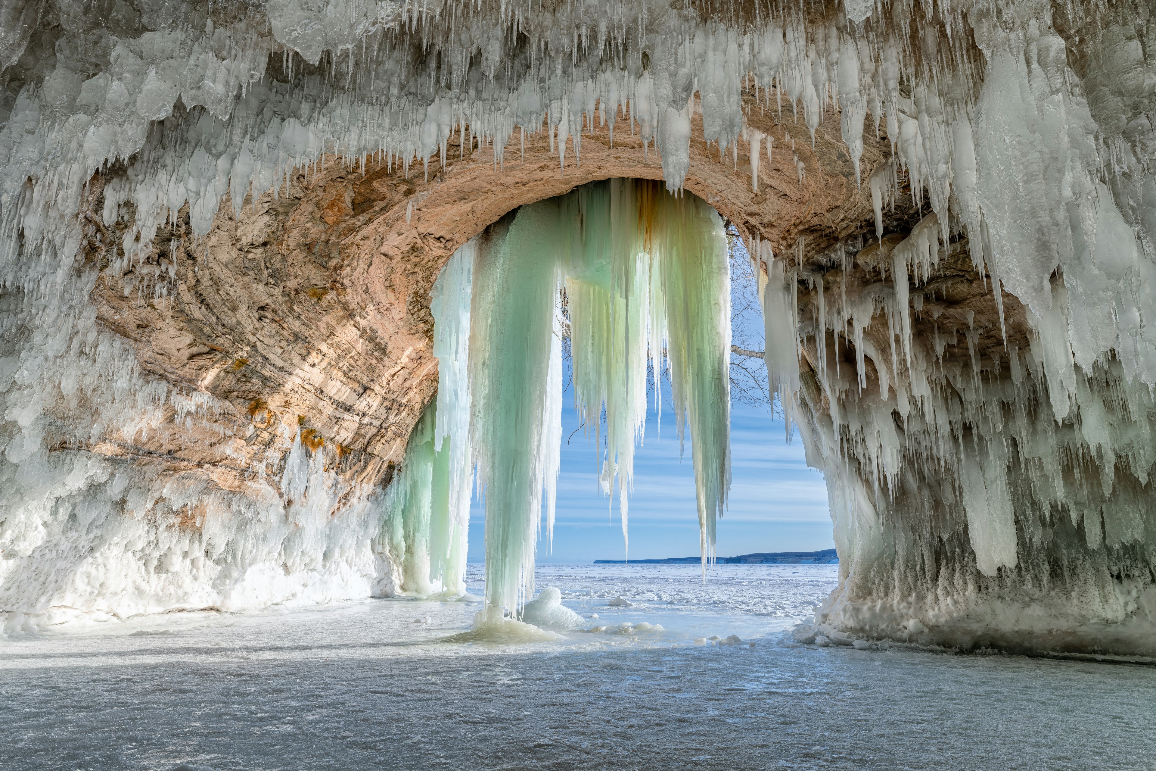 Bright white stalactites framing the mouth of a cave, with a bright blue sky and snowy ground seen through the opening