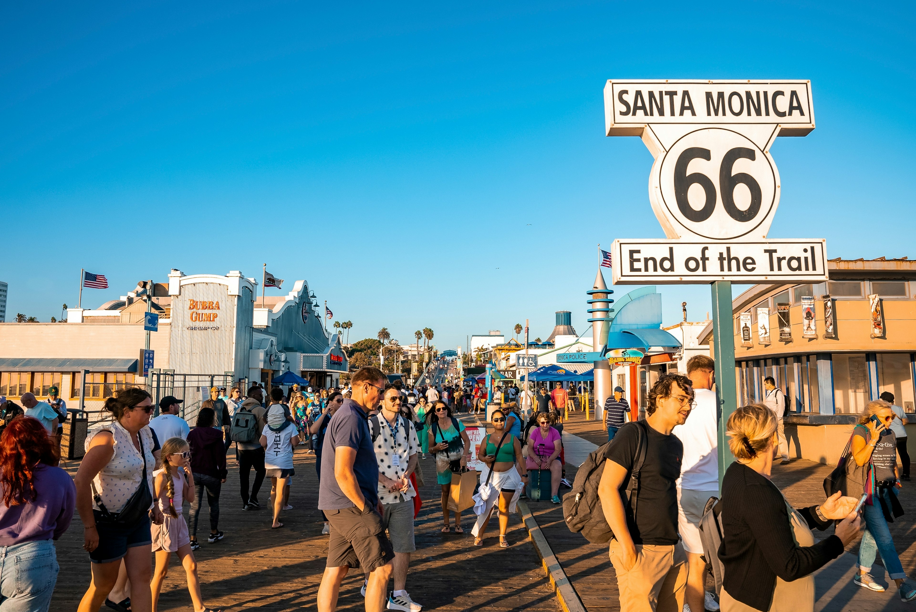 People walking by Santa Monica 66 end of the trail road sign with Pacific Amusement Park and clear blue sky in the background during sunset;