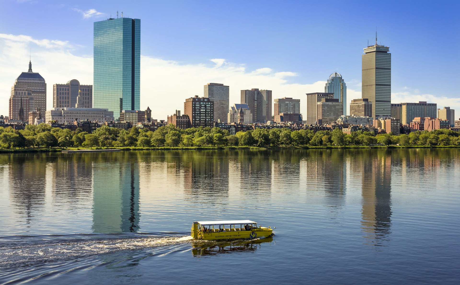 An amphibious boat sails along a river backed by some high-rise city buildings