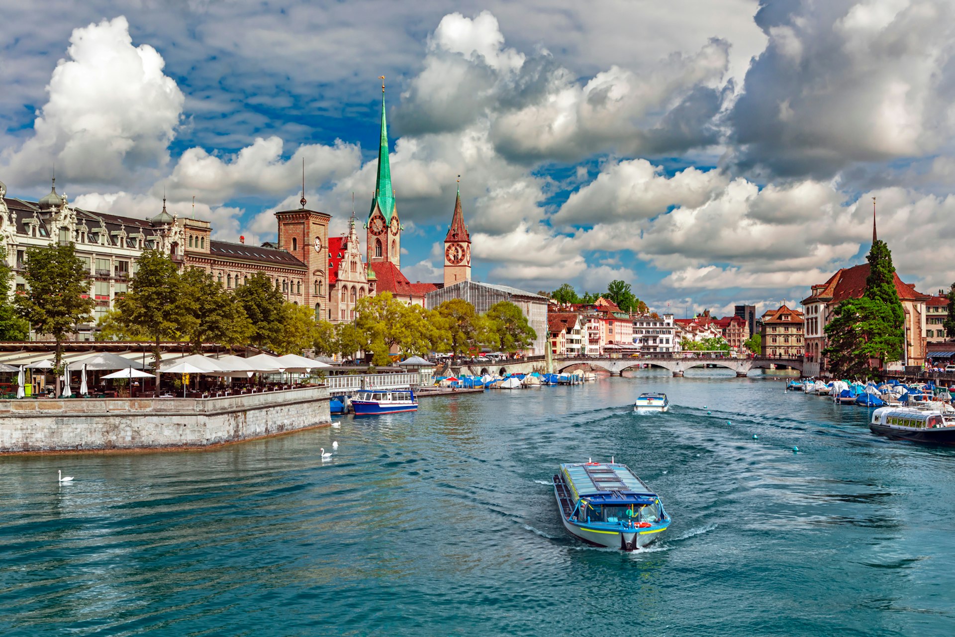 A boat sails along a river in a city. The skyline is dominated by two large church steeples