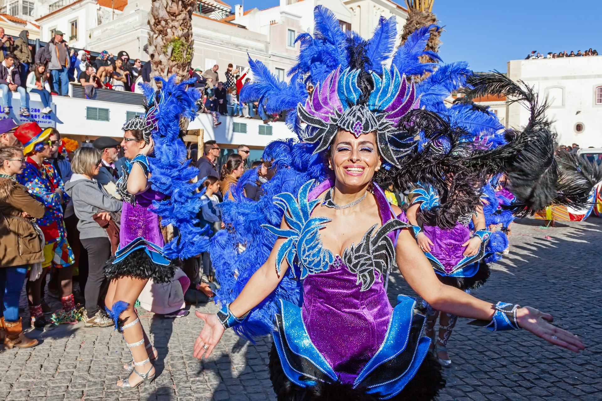 A smiling samba performer in blue-and-purple costume dances in a parade