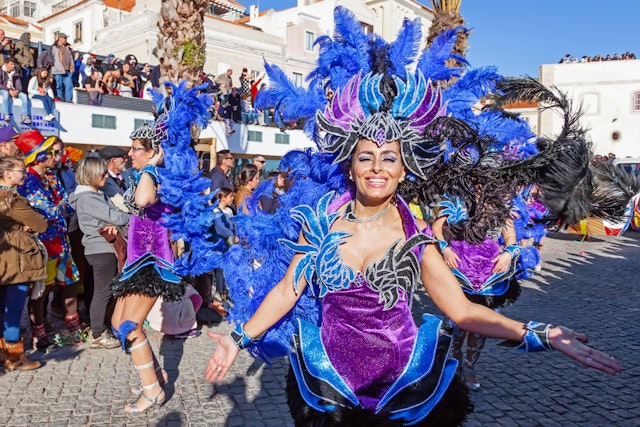 A smiling samba performer in blue-and-purple costume dances in a parade