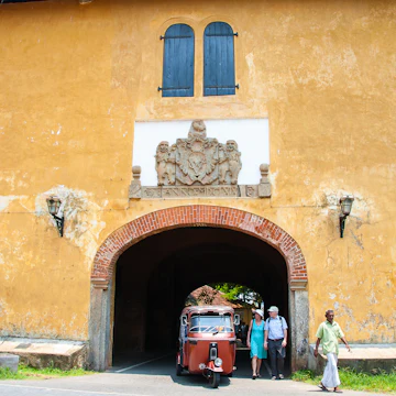 Tuk tuk drives through the entrance of the old Dutch gate with British Coat of Arms above.