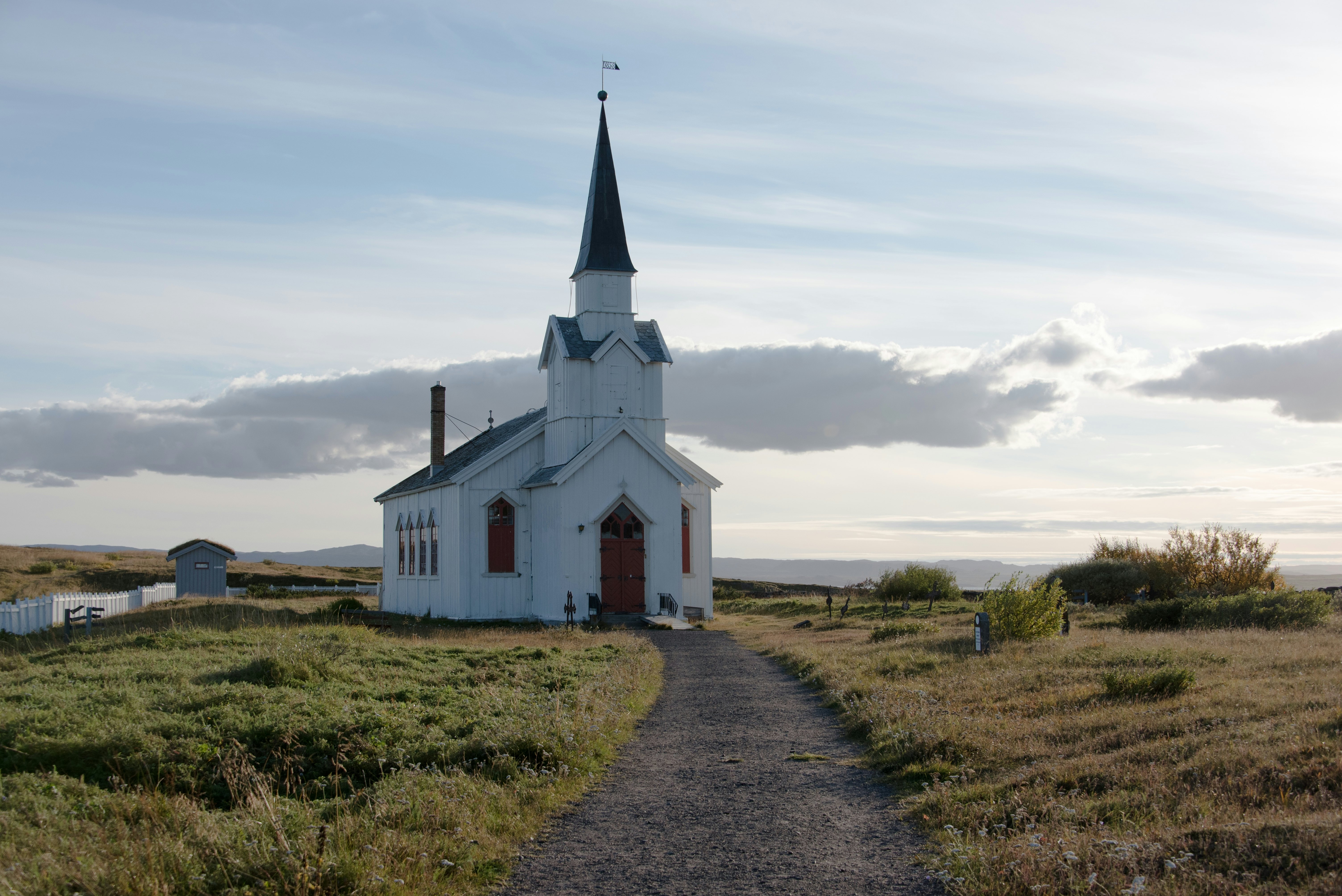 A small, plain, white church with spire at the end of a dirt path surrounded by low, autumnal grass on a partly sunny day.