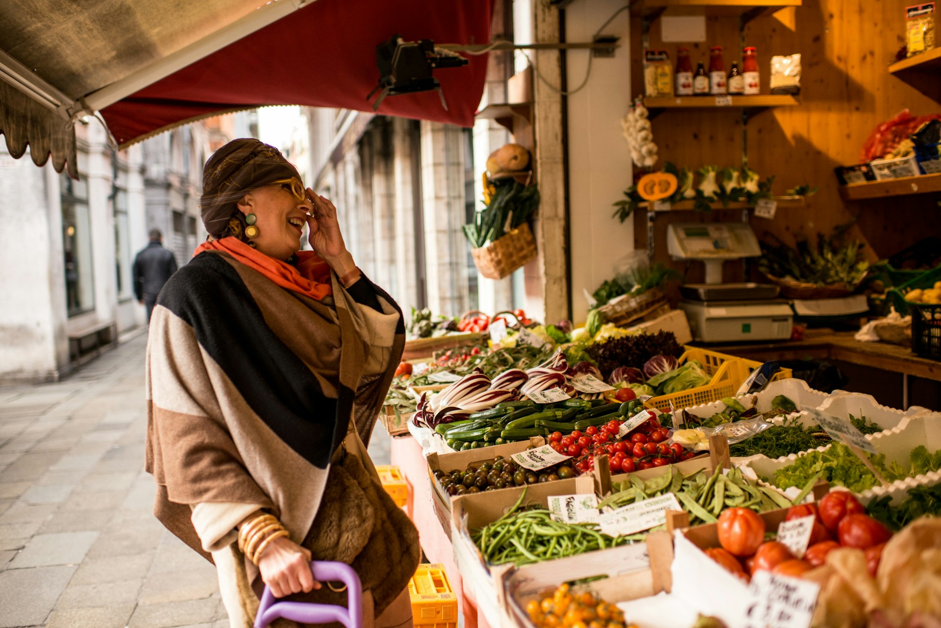 An older woman, dressed in winter clothes and wearing a turban, is smiling at the grocery market stall in Venice.
