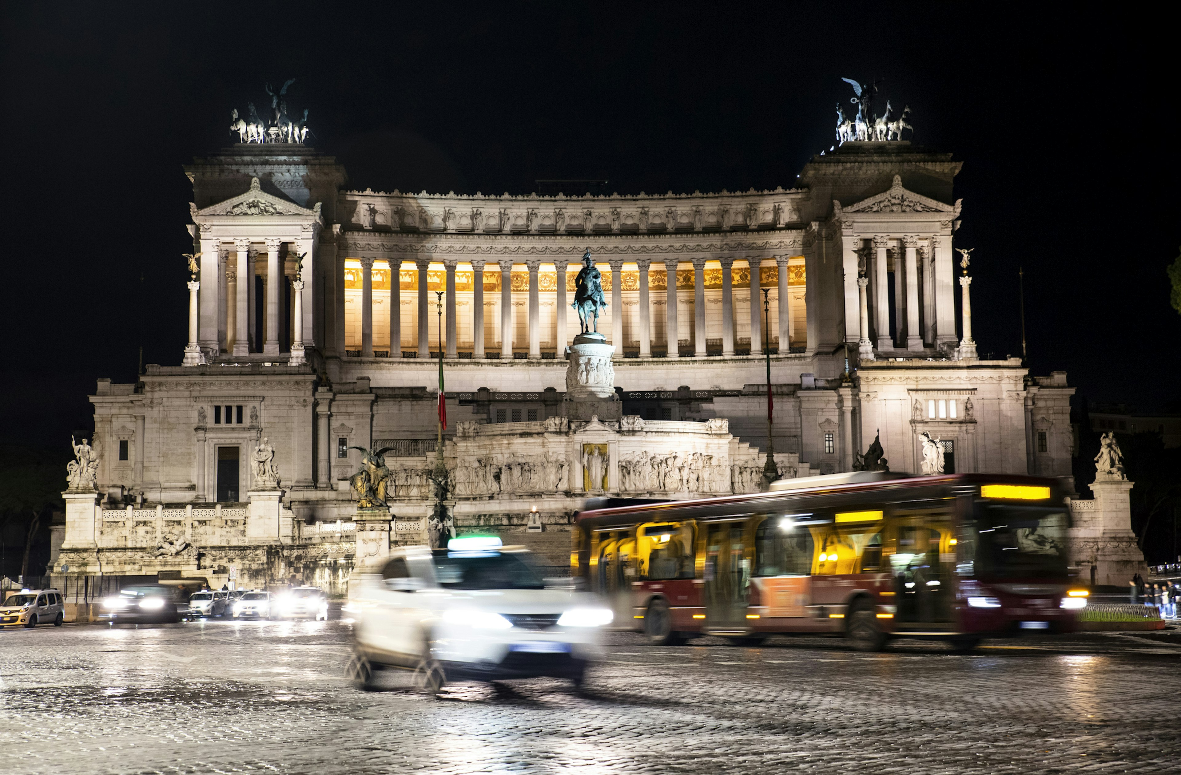 A taxi and a bus pass a huge, grand building lit up at night in Rome