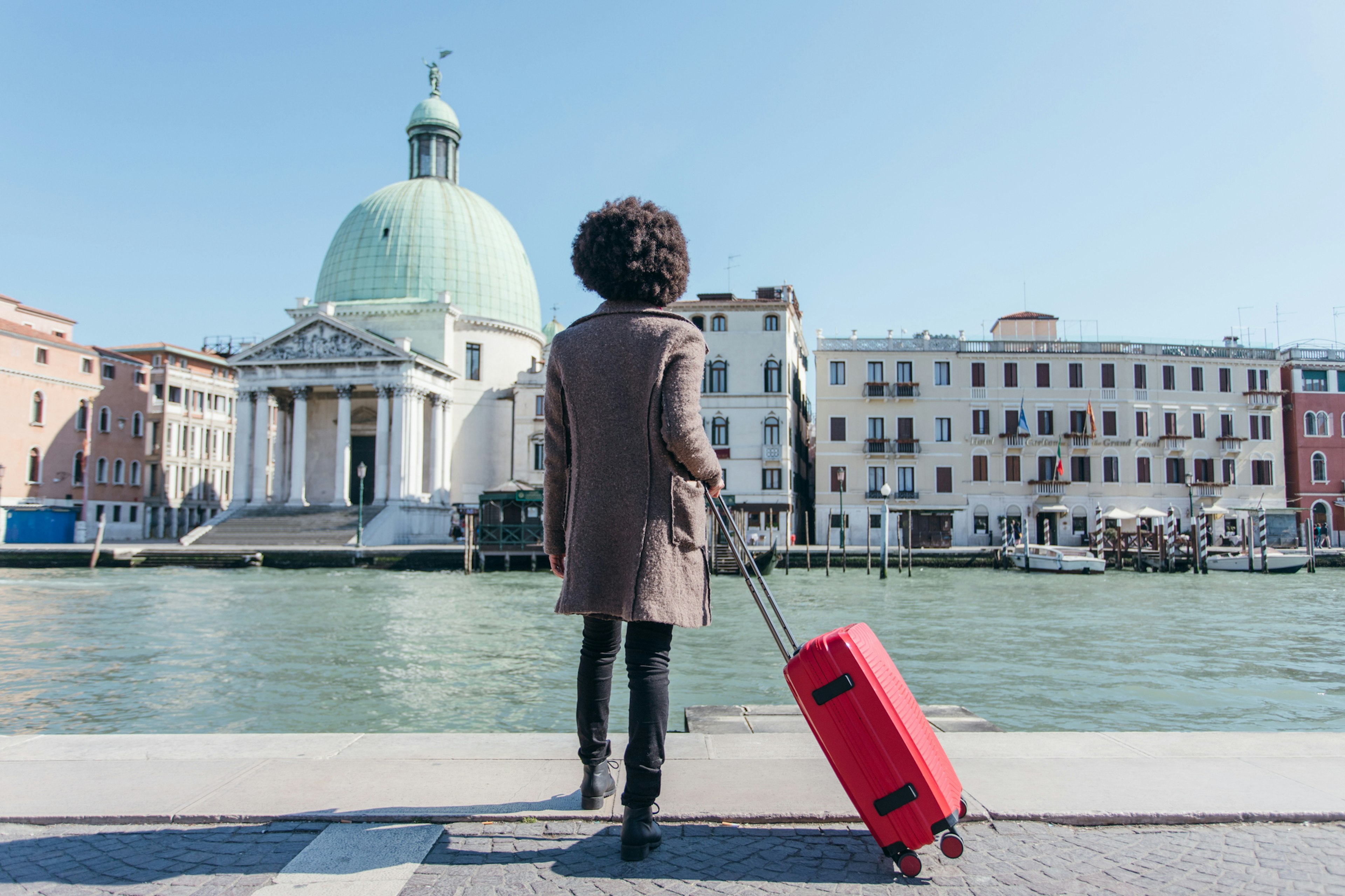 A woman standing on the side of a canal with a suitcase in Venice