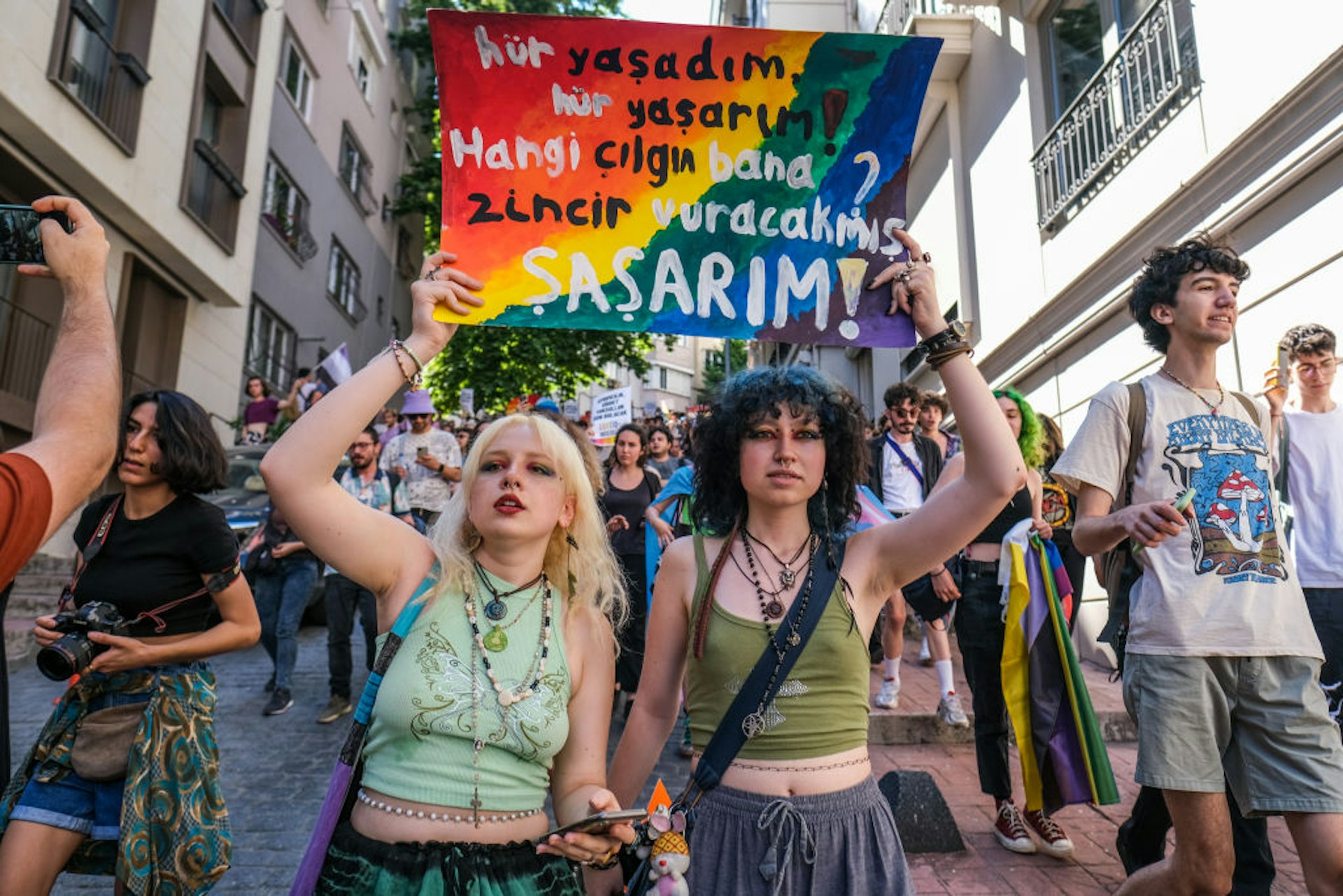 Two women holding rainbow signs protest for the rights of LGBTQ people in a crowded street. 