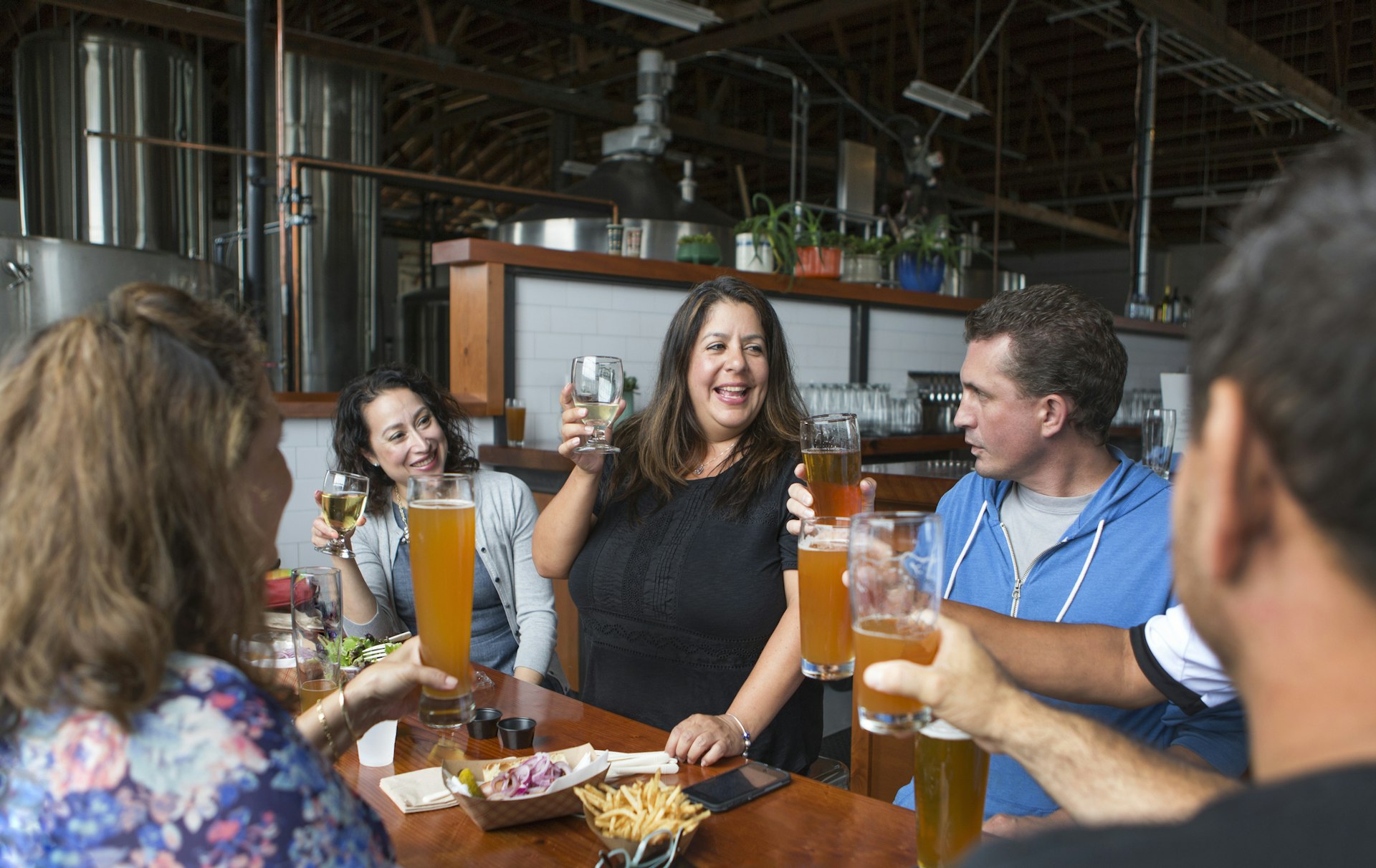 People drink beer and wine in a brewery, with an out-of-focus bar and stainless steel beer manufacturing equipment in the background. 